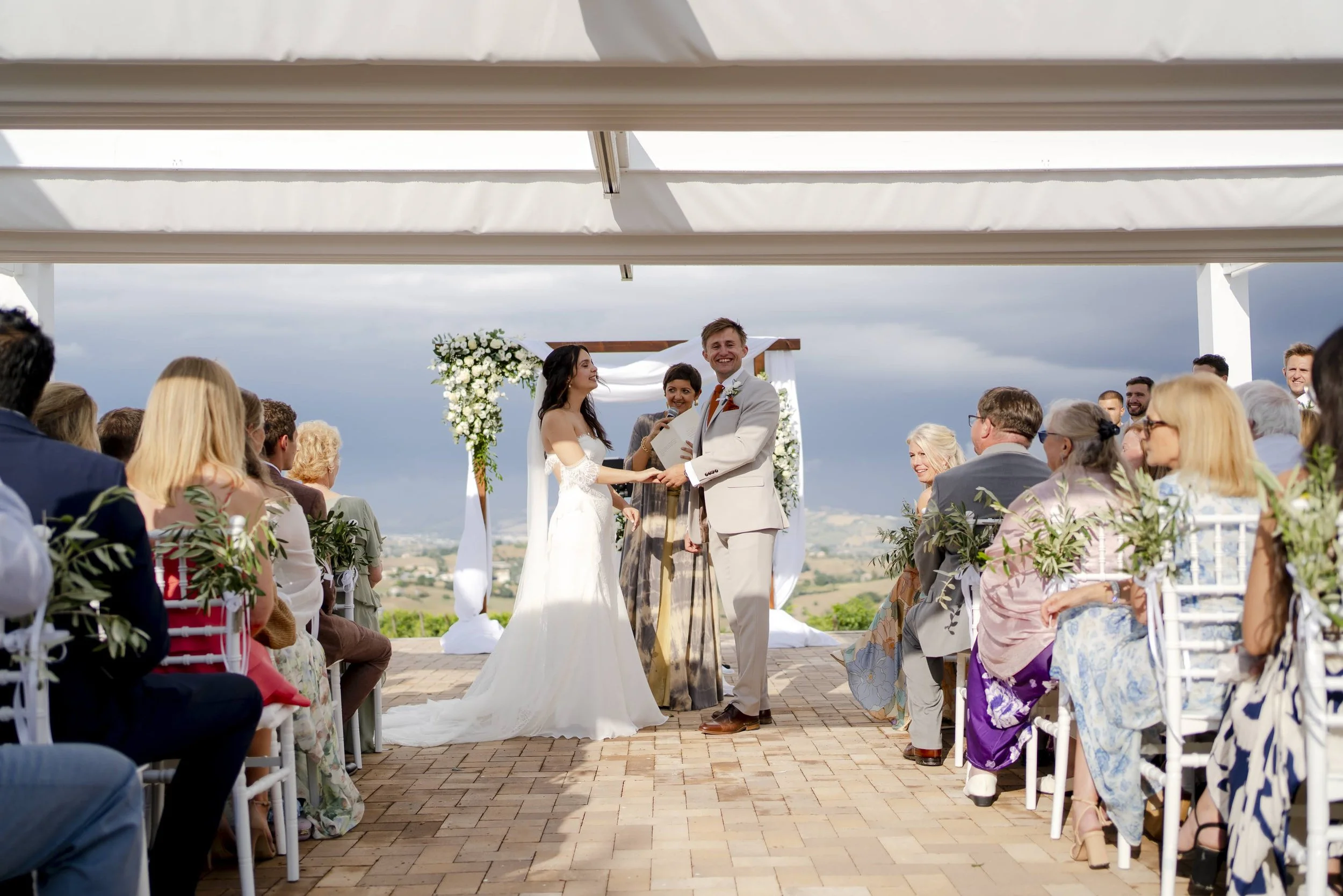 A wedding ceremony outdoors with the bride and groom exchanging vows under a decorated arch, surrounded by seated guests on a brick patio with a scenic countryside background.