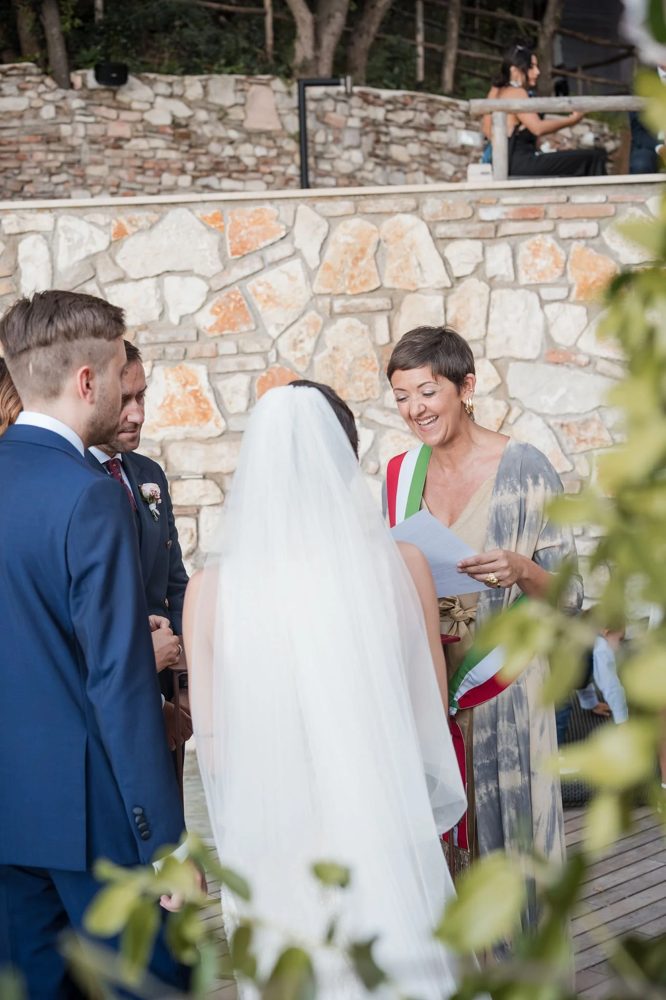 A wedding ceremony outdoors with a woman officiant wearing a sash, speaking to the bride and groom in front of a stone wall, with some greenery in the foreground.