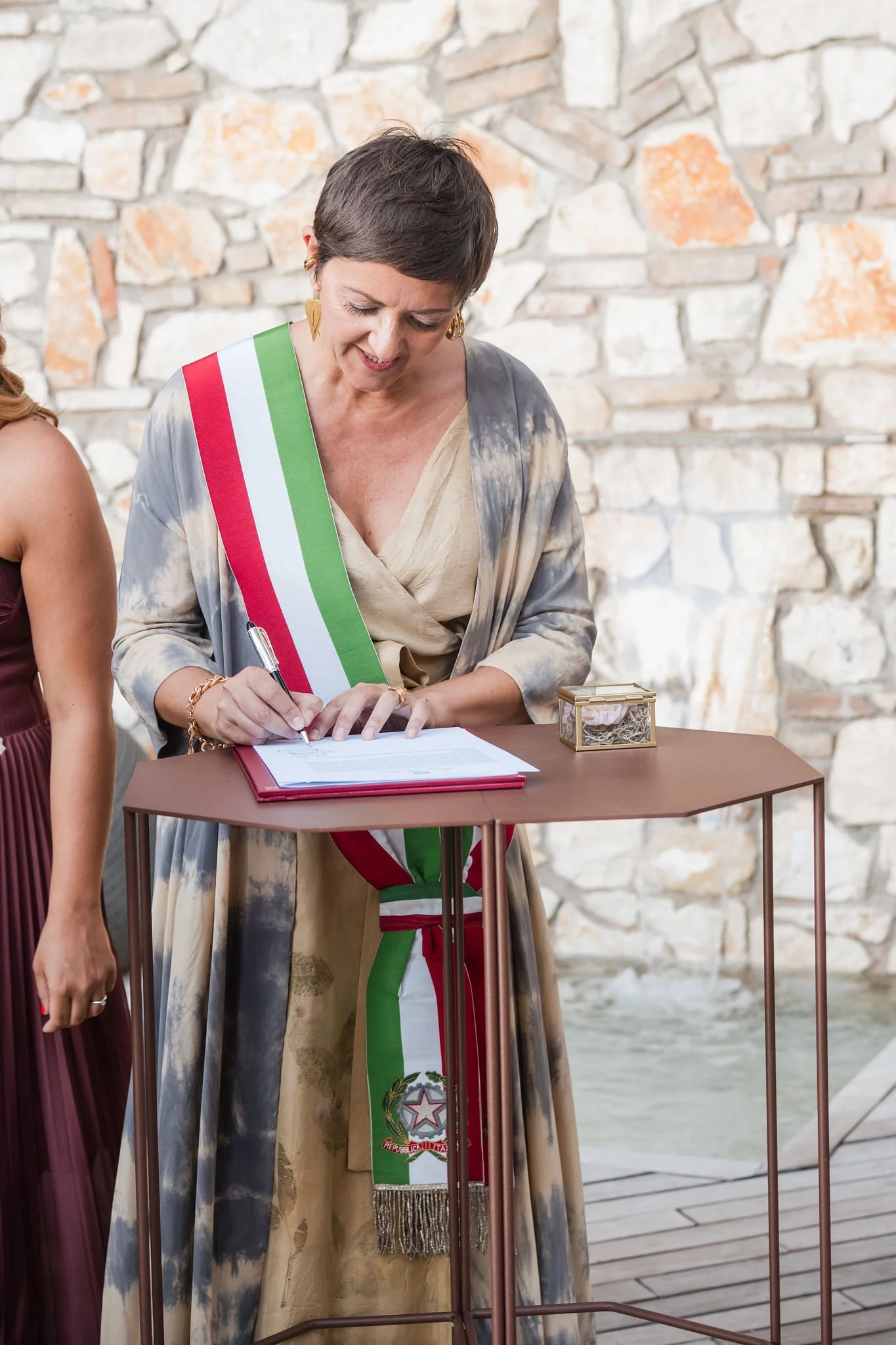 A woman wearing a sash with the colors of the Italian flag signing a document at a wooden table. She is wearing a patterned dress with a beige top and a colorful wrap. There is a small decorative box on the table, and a stone wall in the background.
