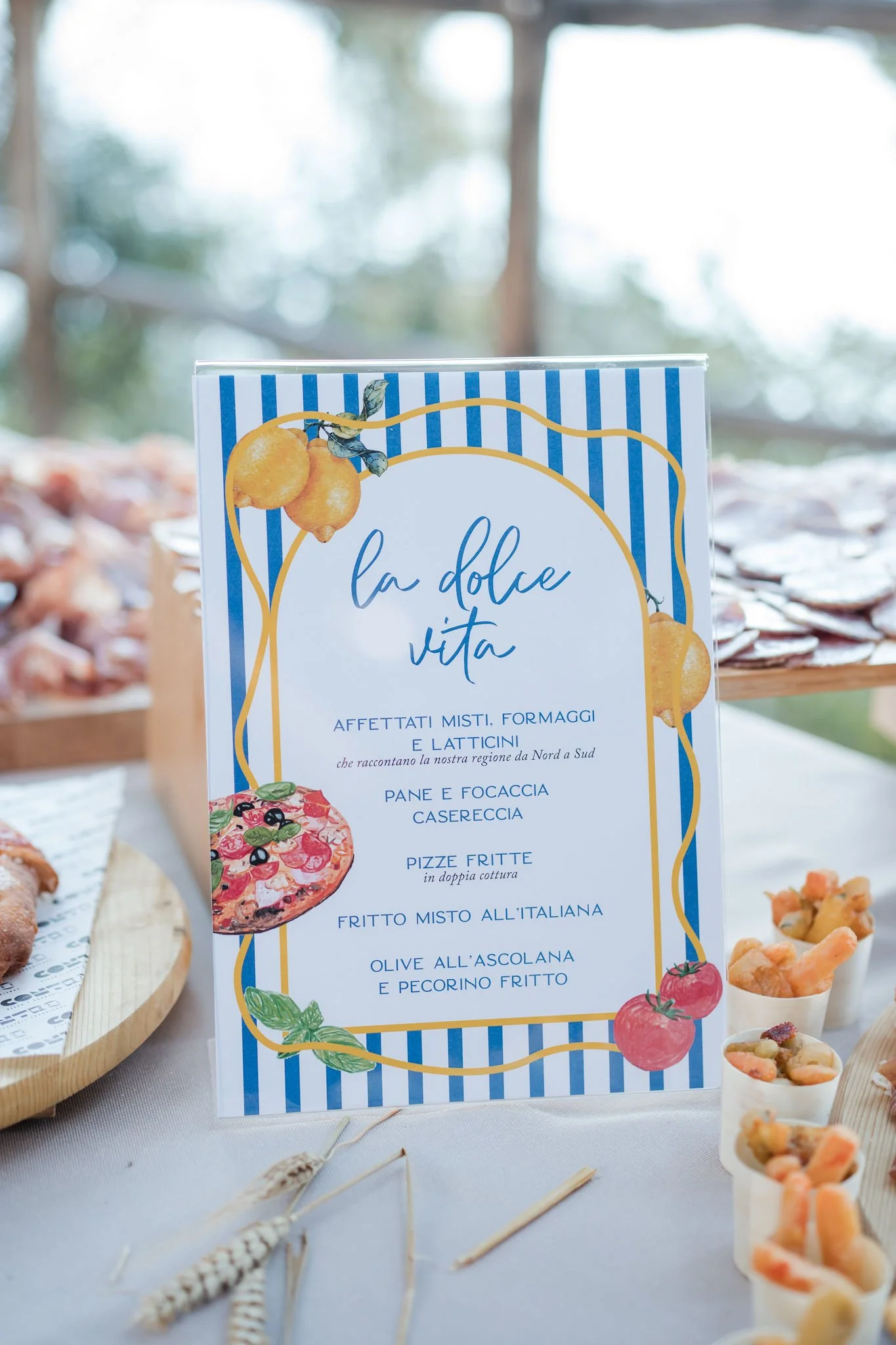 Menu sign titled 'La Dolce Vita' with Italian dishes list, decorated with illustrations of lemons, tomatoes, and basil, placed on a table with assorted small dishes and appetizers in the background.