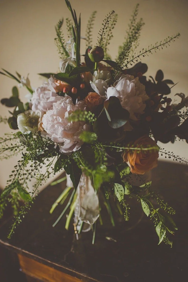 A bouquet of mixed flowers including pink peonies, orange roses, and greenery arranged in a vase, placed on a dark wooden surface.