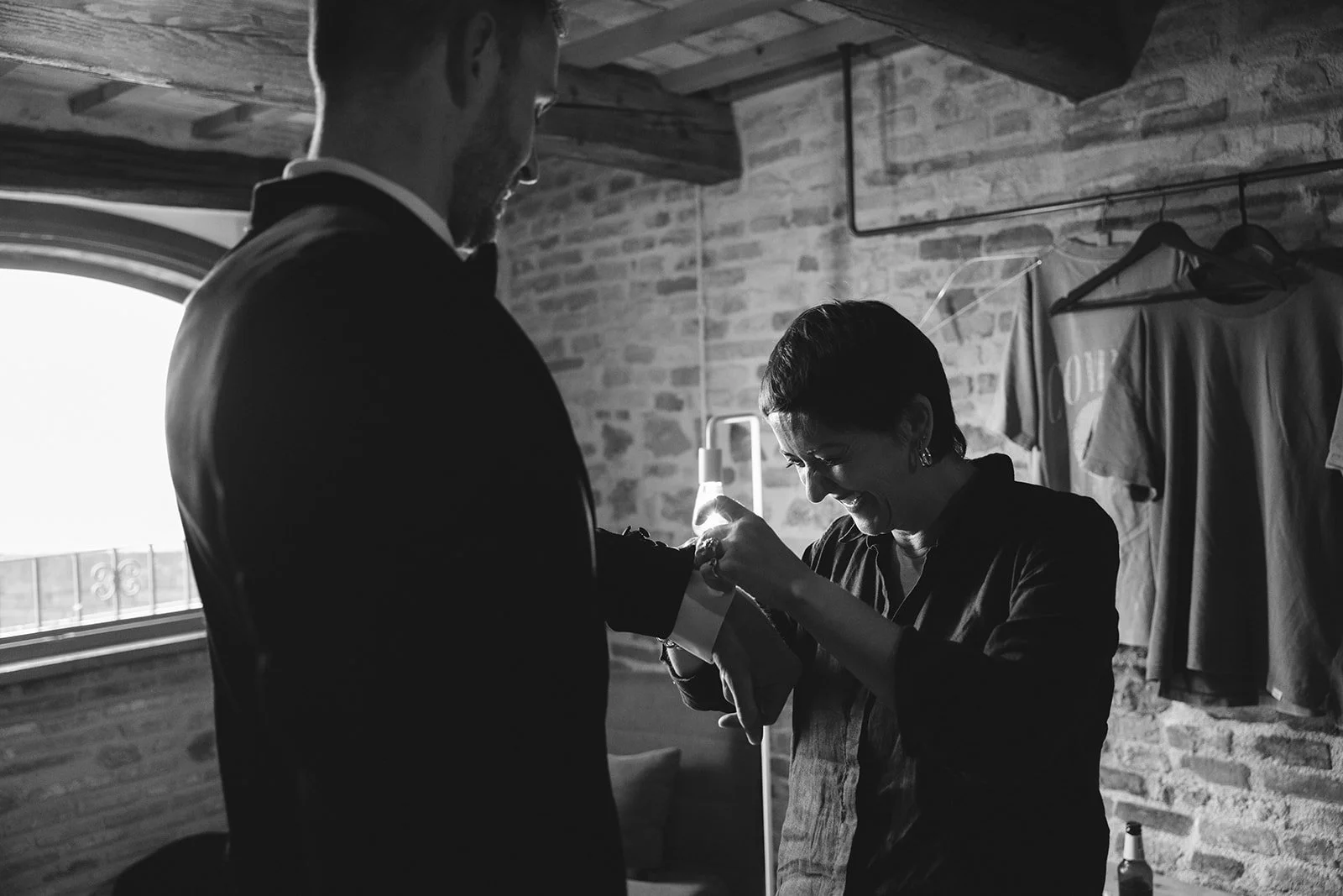 A woman joyfully gets an accessory placed on her wrist by a man in a suit, in a rustic indoor space with brick walls and hanging clothes.