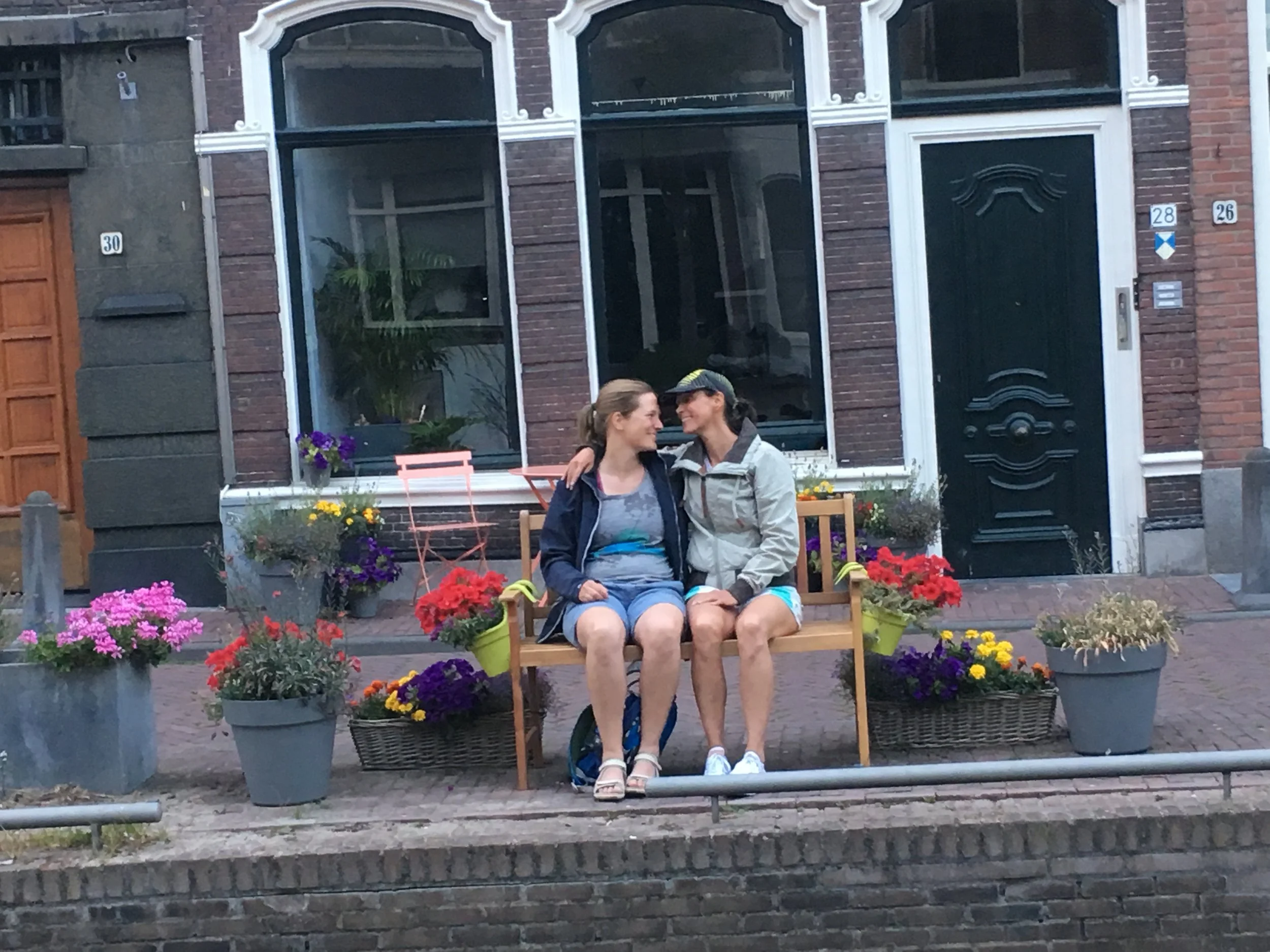Two women sitting closely on a wooden bench, smiling at each other, surrounded by colorful potted flowers on a city sidewalk.