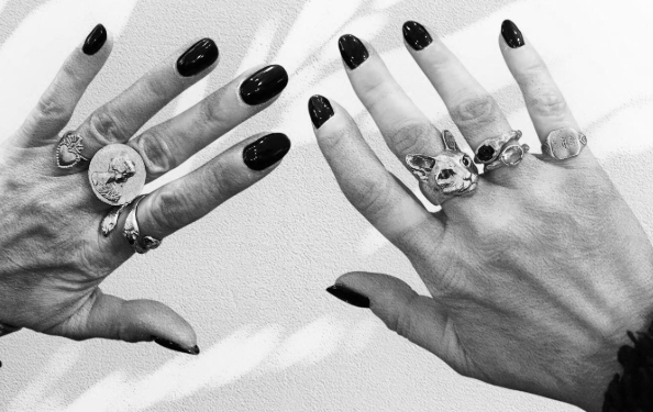 Two hands with black nail polish, each wearing multiple rings with various designs, including animals and intricate patterns, positioned against a plain background.