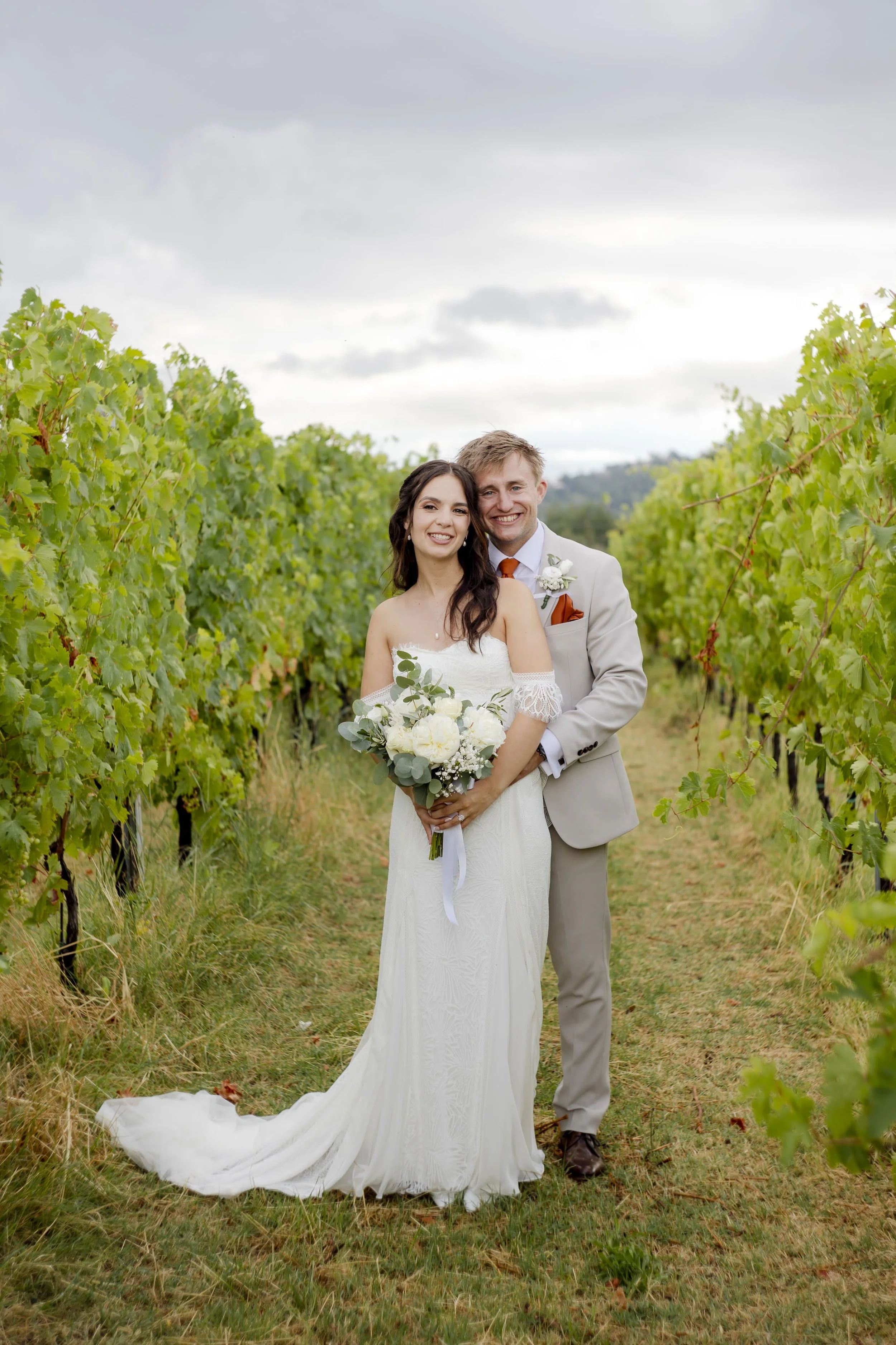 A bride and groom standing together in a vineyard, smiling, with the bride holding a bouquet of white flowers and greenery.