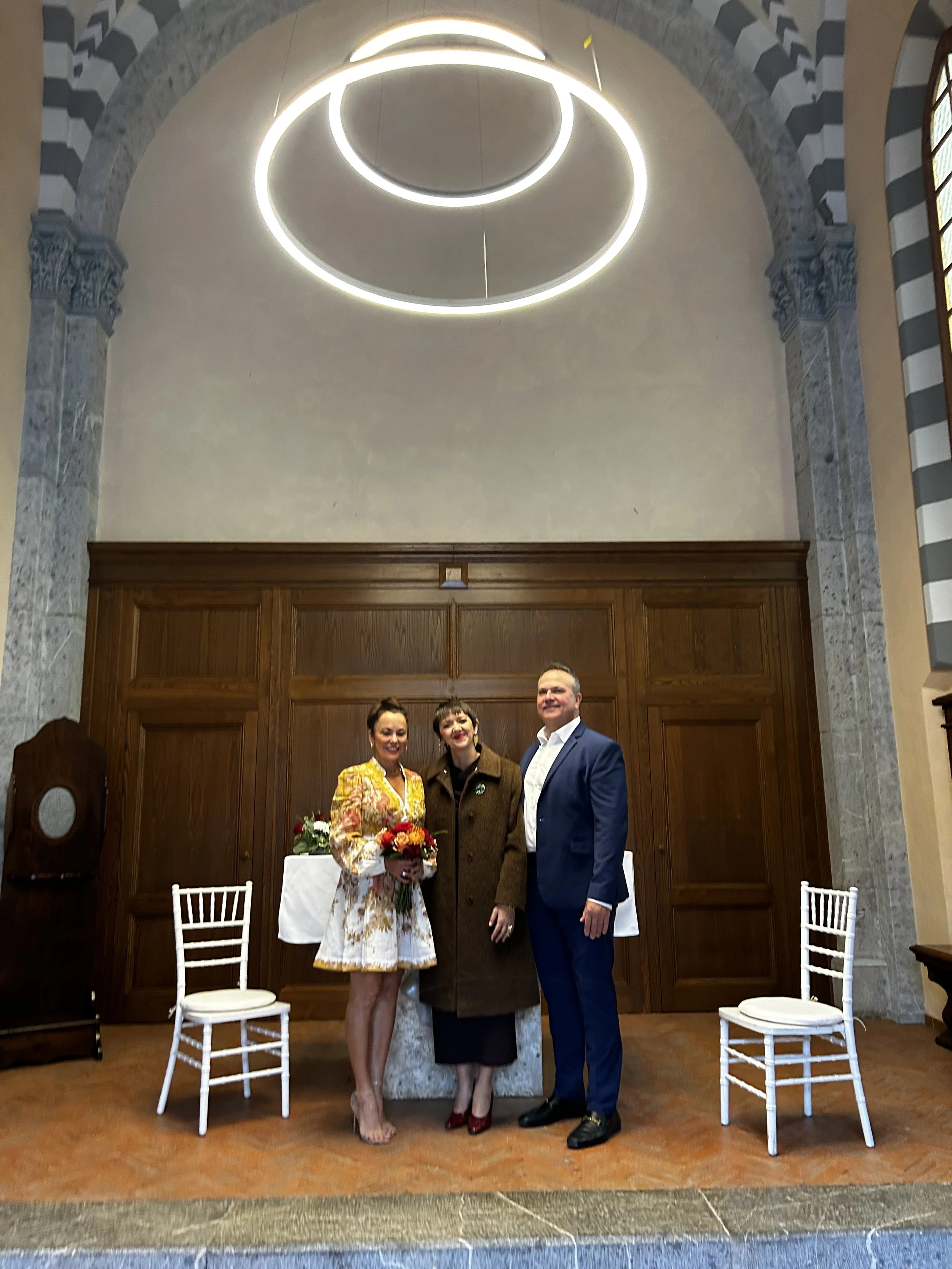 Three people standing together inside a church, a woman in a floral dress holding a bouquet, a woman in a brown coat, and a man in a blue suit, with empty white chairs on each side.