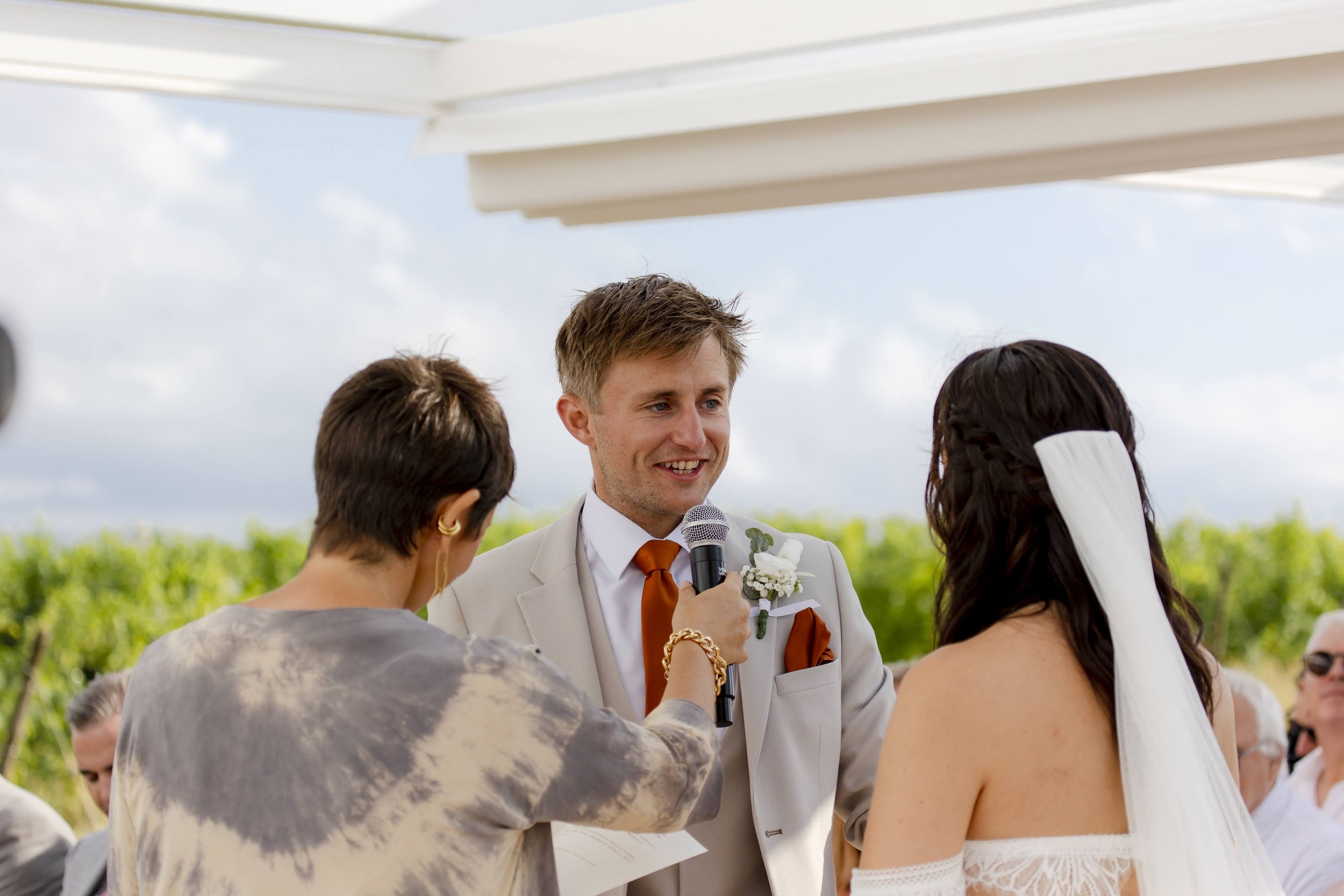 A man in a beige suit and orange tie is smiling at a woman with long dark hair and a white veil during a wedding ceremony outdoors. A woman with short brown hair and gold earrings holds a microphone near the man, with guests seated behind them and gr