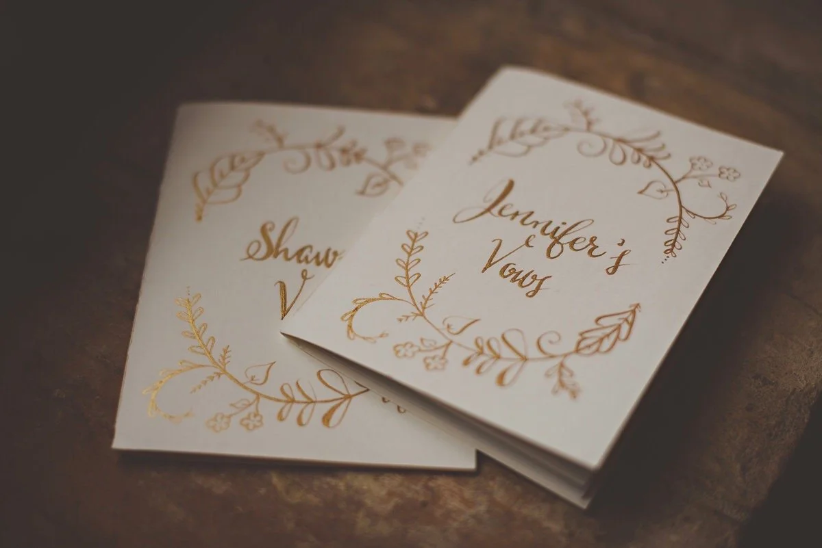 Wedding vows booklet with gold floral designs on white paper, titled "Jennifer's Vows" in cursive, resting on a wooden surface.