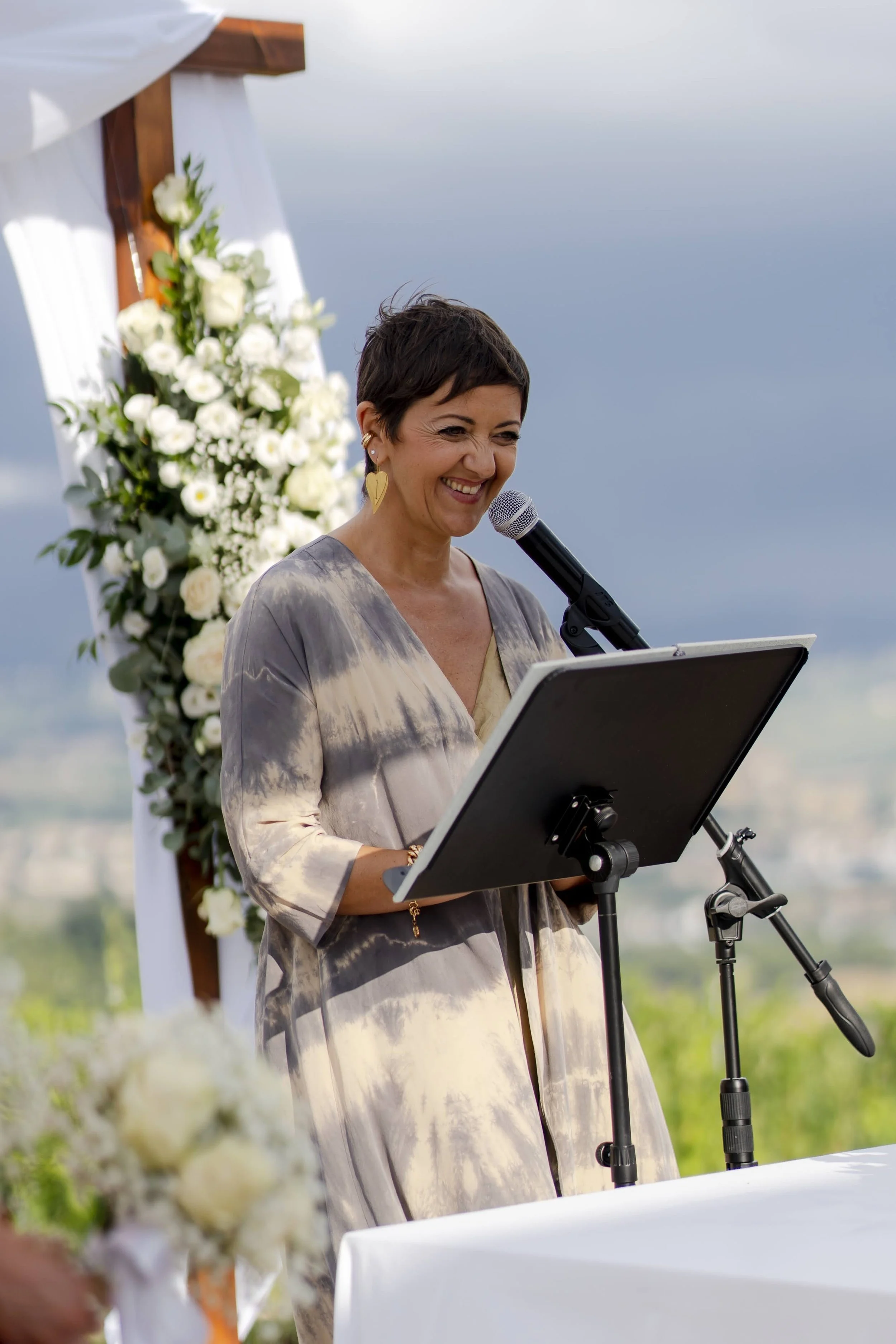 A woman giving a speech at an outdoor wedding ceremony, standing behind a podium with a microphone, with a floral arrangement and a wedding arch in the background.