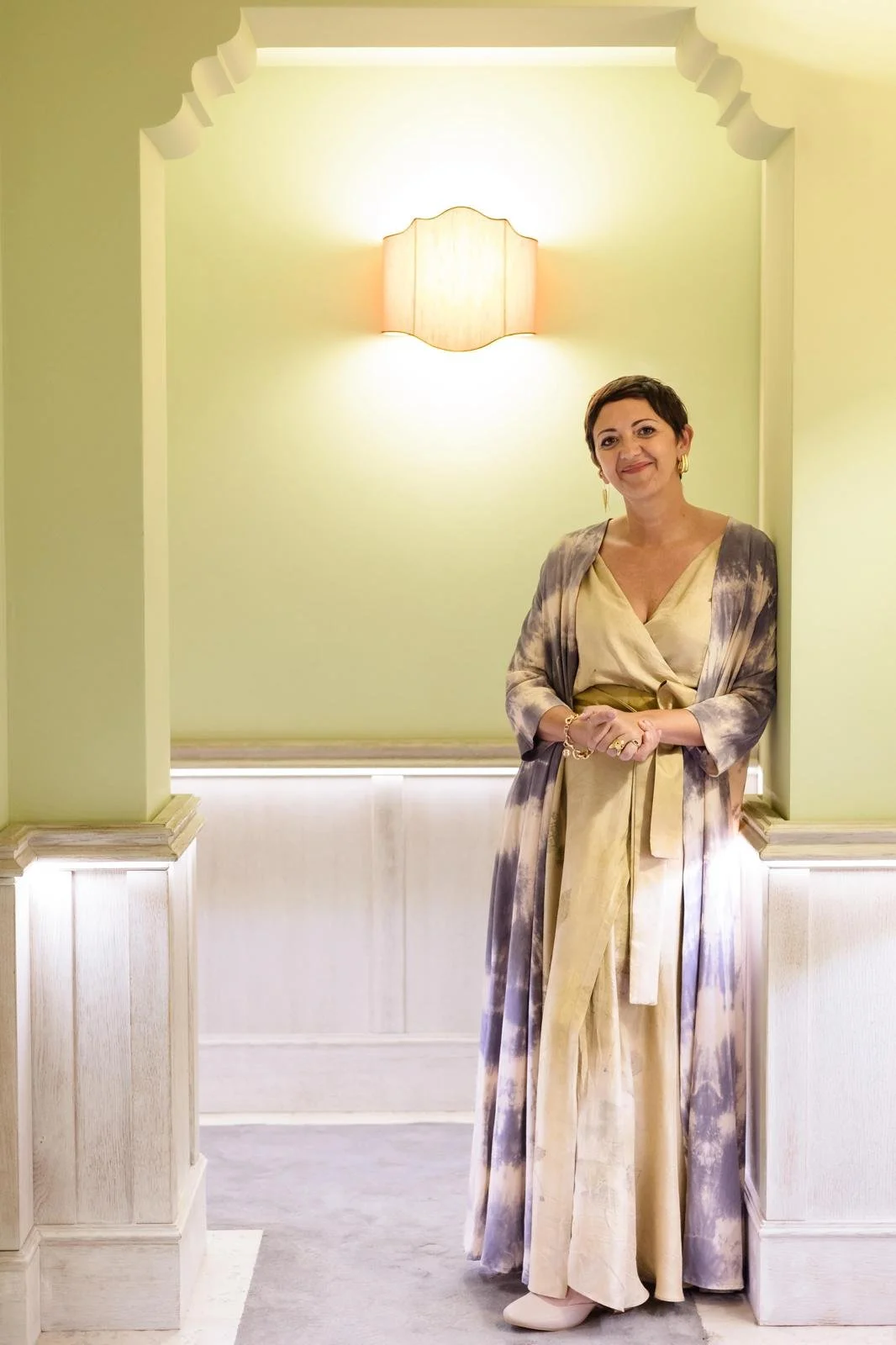 A woman standing in a doorway with a soft smile, wearing a flowing cream dress and a tie-dye patterned cardigan, inside a room with light green walls and decorative trim, illuminated by a wall sconce light.