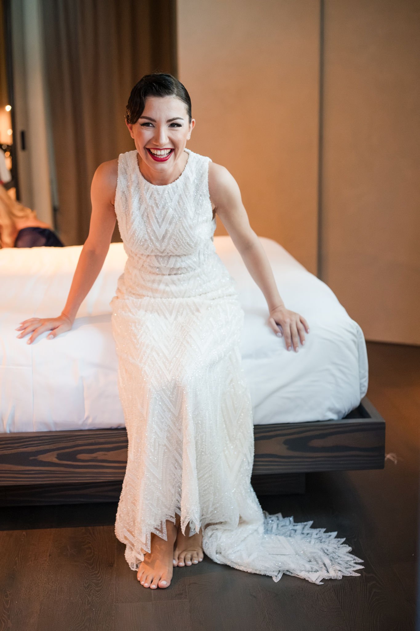A woman in a white, beaded, sleeveless wedding dress sitting on the edge of a bed, smiling happily at the camera.