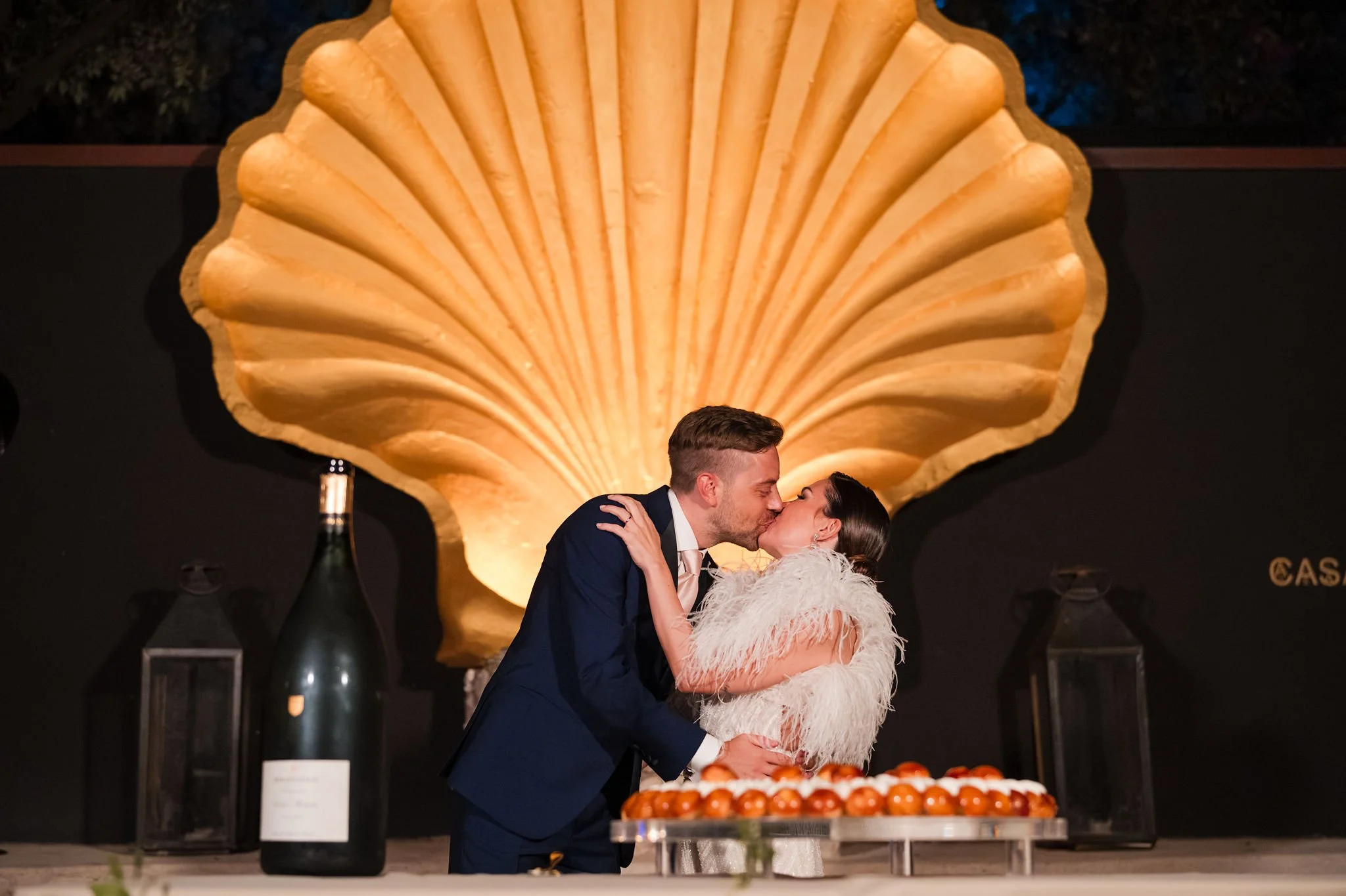 A newlywed couple in wedding attire sharing a kiss at their wedding reception, with a large decorative seashell backdrop behind them and a table with a cake and champagne nearby.