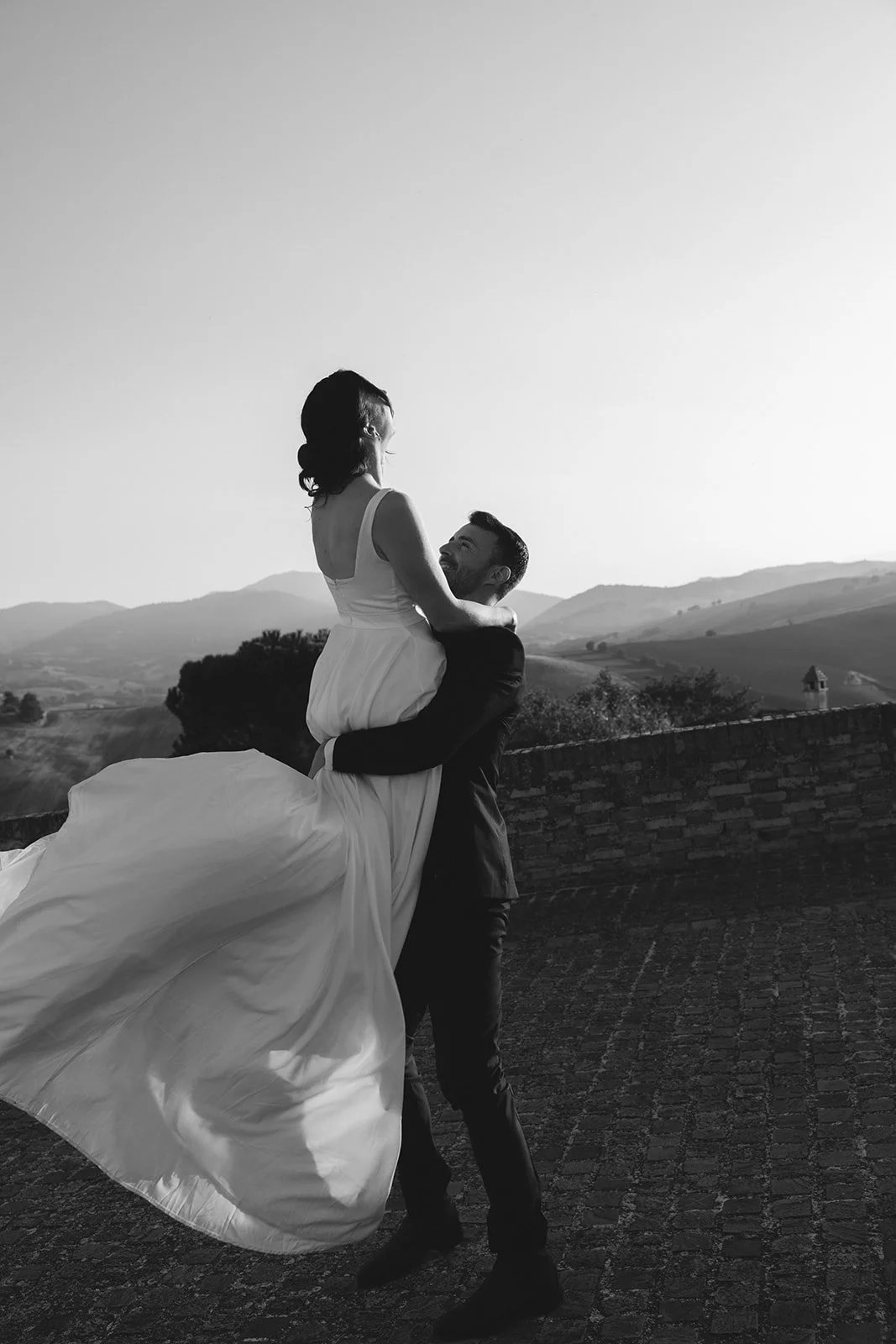 A black-and-white photo of a bride and groom outdoors, with the groom lifting the bride, both smiling and looking at each other, mountains and open sky in the background.