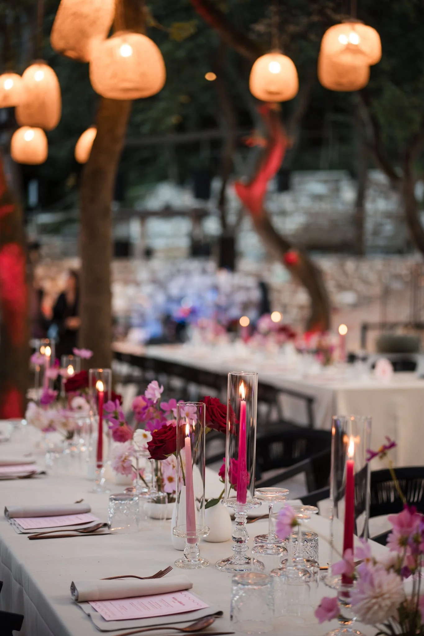 Elegant outdoor dinner table decorated with pink and white flowers, tall candles in glass holders, set in a wooded area with hanging paper lanterns.