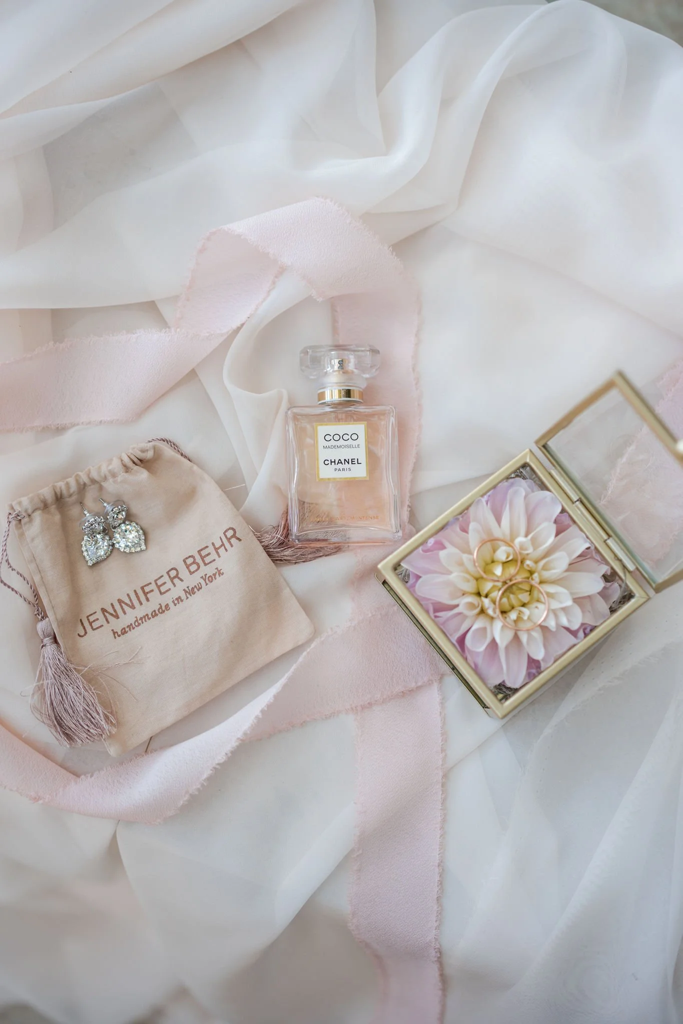 A Chanel Coco Mademoiselle perfume bottle, a pair of diamond butterfly earrings on a Jennifer Behr pouch, a pink ribbon, and a jewelry box with a flower inside on a soft, pale fabric background.