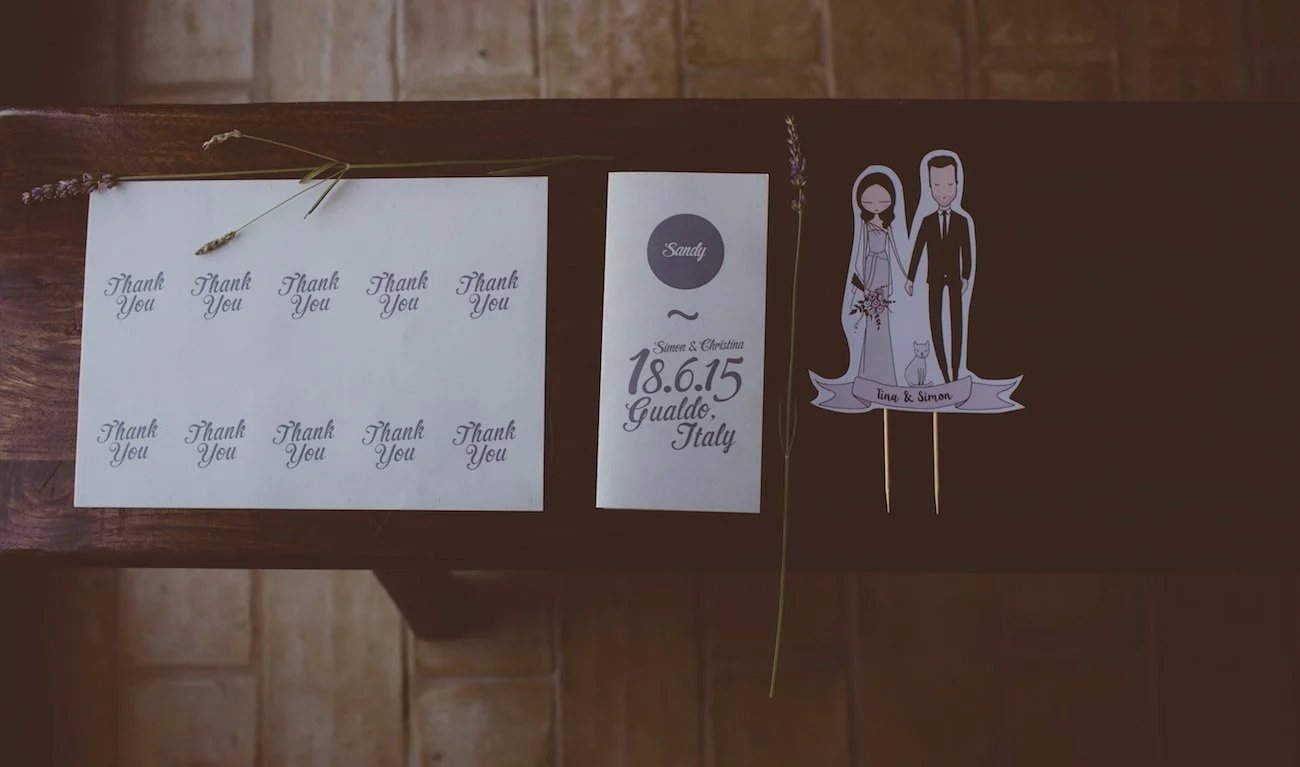 Wedding thank you notes, an event card with the names Simone and Christina, and a paper cutout of a bride and groom with the names Tina and Simon, on a wooden table with some lavender sprigs.