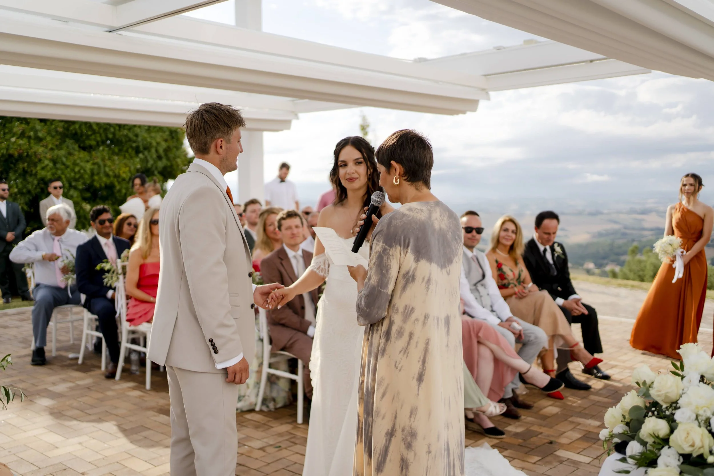 A wedding ceremony outdoors with a bride and groom holding hands and listening to an officiant reading from a paper, surrounded by seated guests and a scenic landscape in the background.