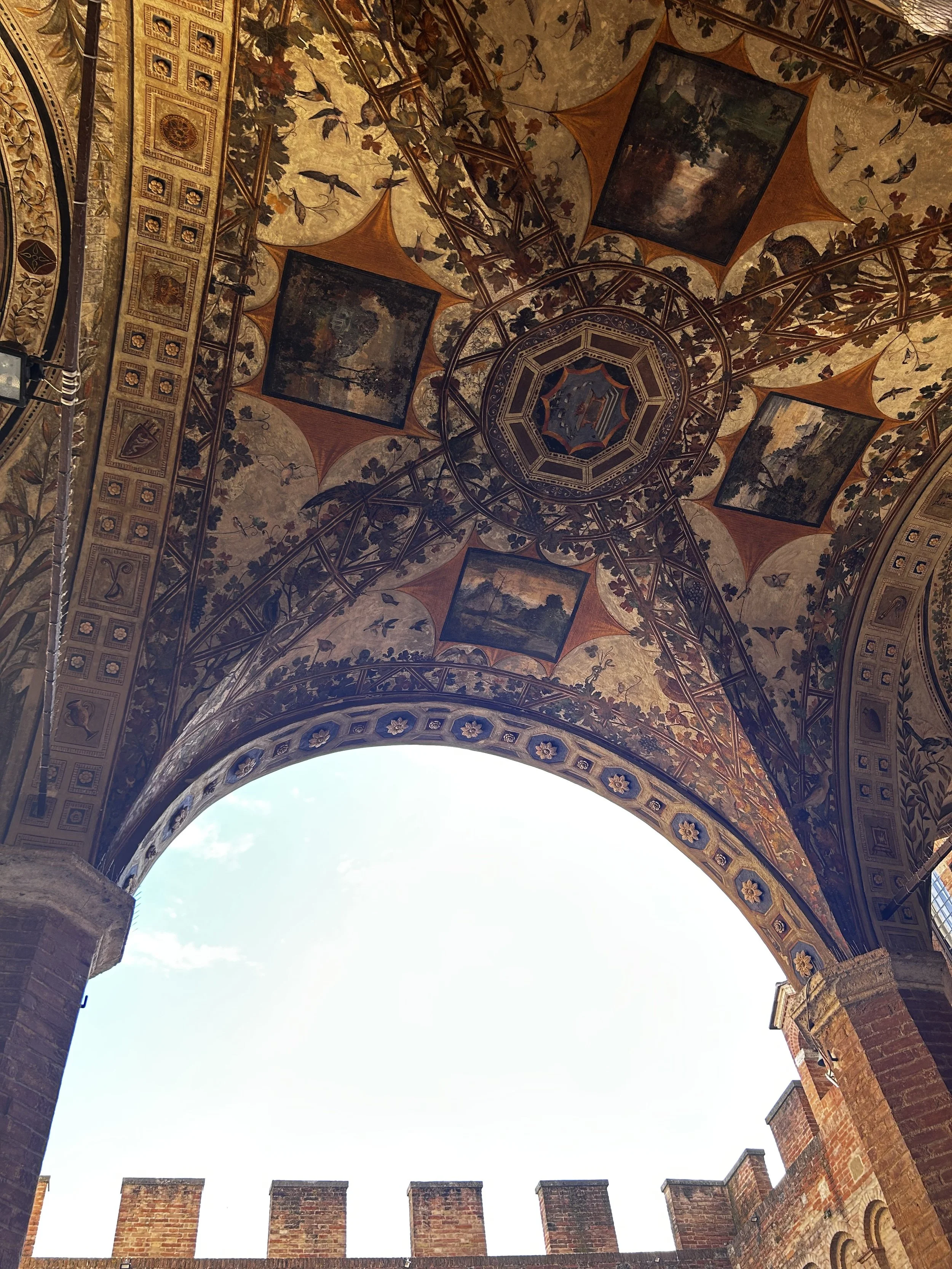 Ornately decorated arched ceiling with paintings and intricate patterns, viewed from below with blue sky visible through the arch.