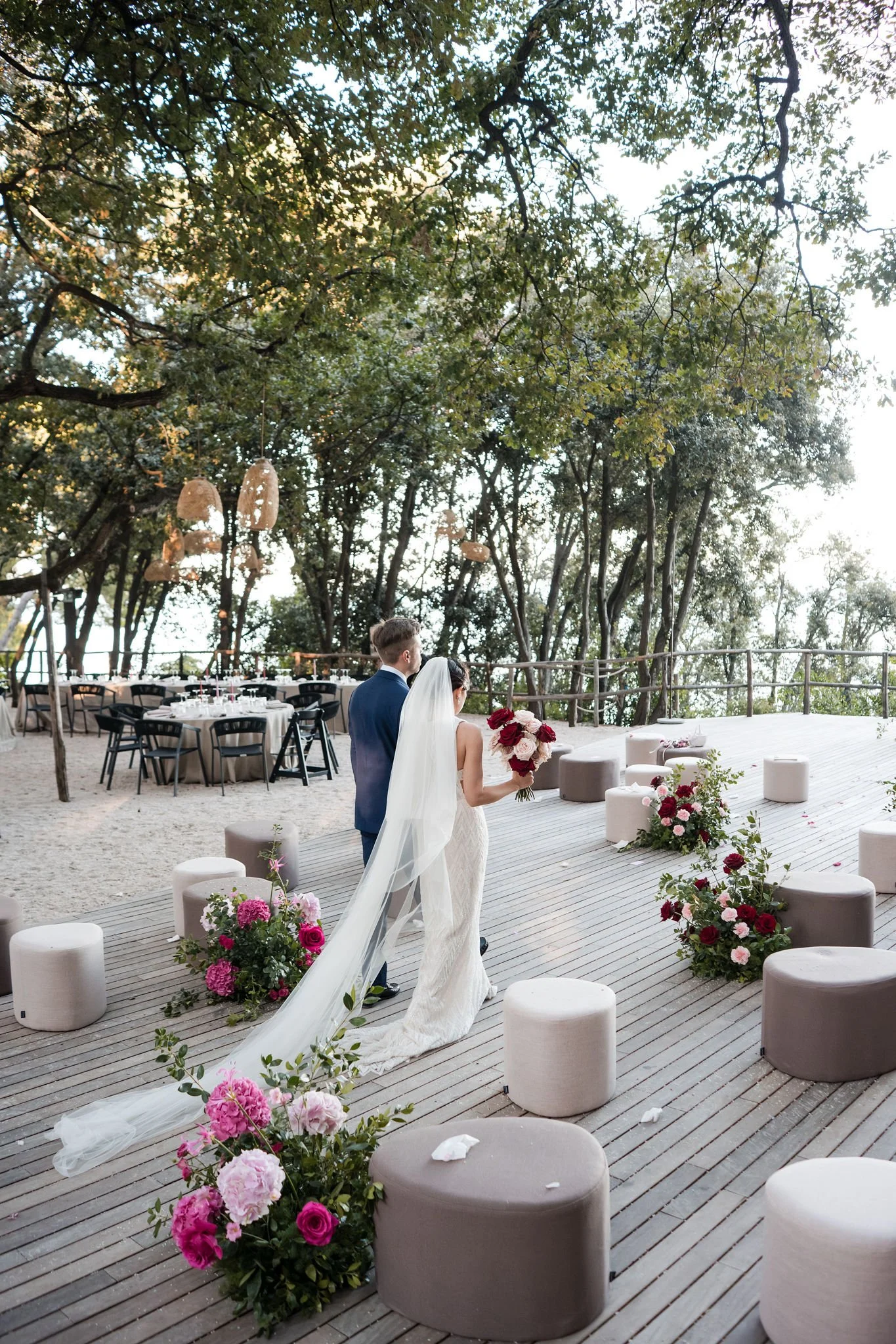 Bride and groom walking down an outdoor wedding aisle decorated with pink and red flowers, under a canopy of trees.