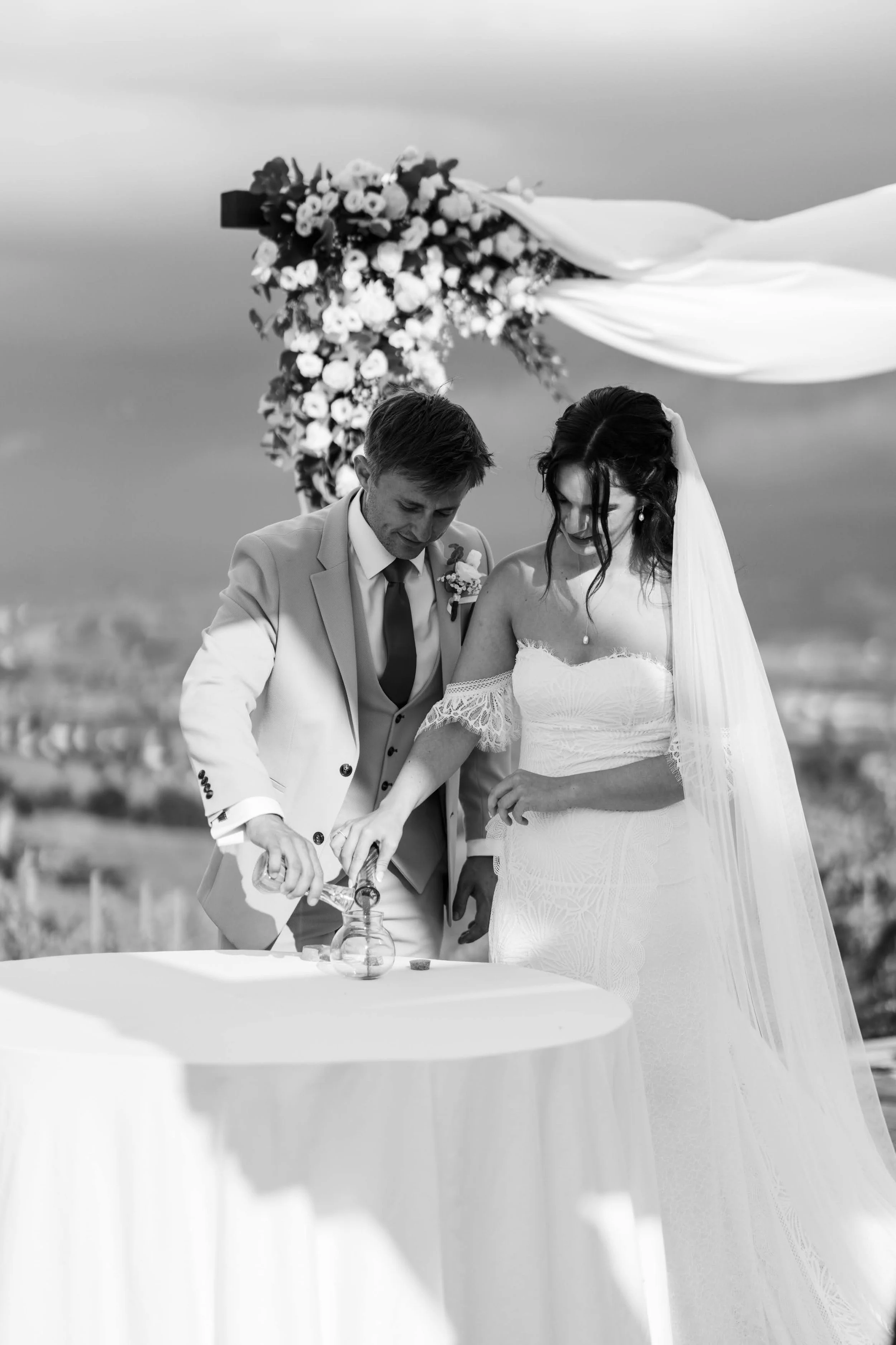 Black-and-white photo of a couple at their wedding ceremony outdoors, with the groom pouring a liquid from a bottle into a glass on a table, and the bride standing beside him. There is a floral arrangement and flowing fabric overhead.