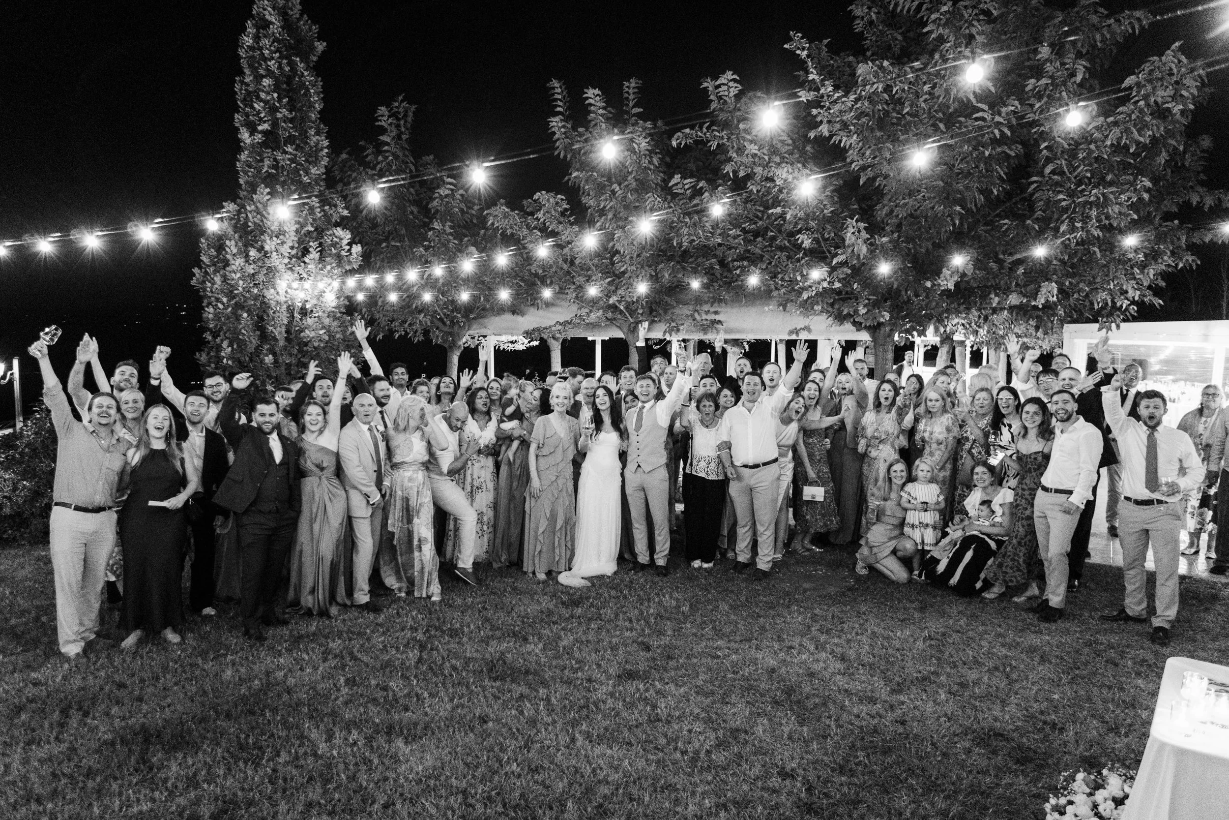 A large group of people gathered outdoors at night, celebrating under string lights with trees in the background.