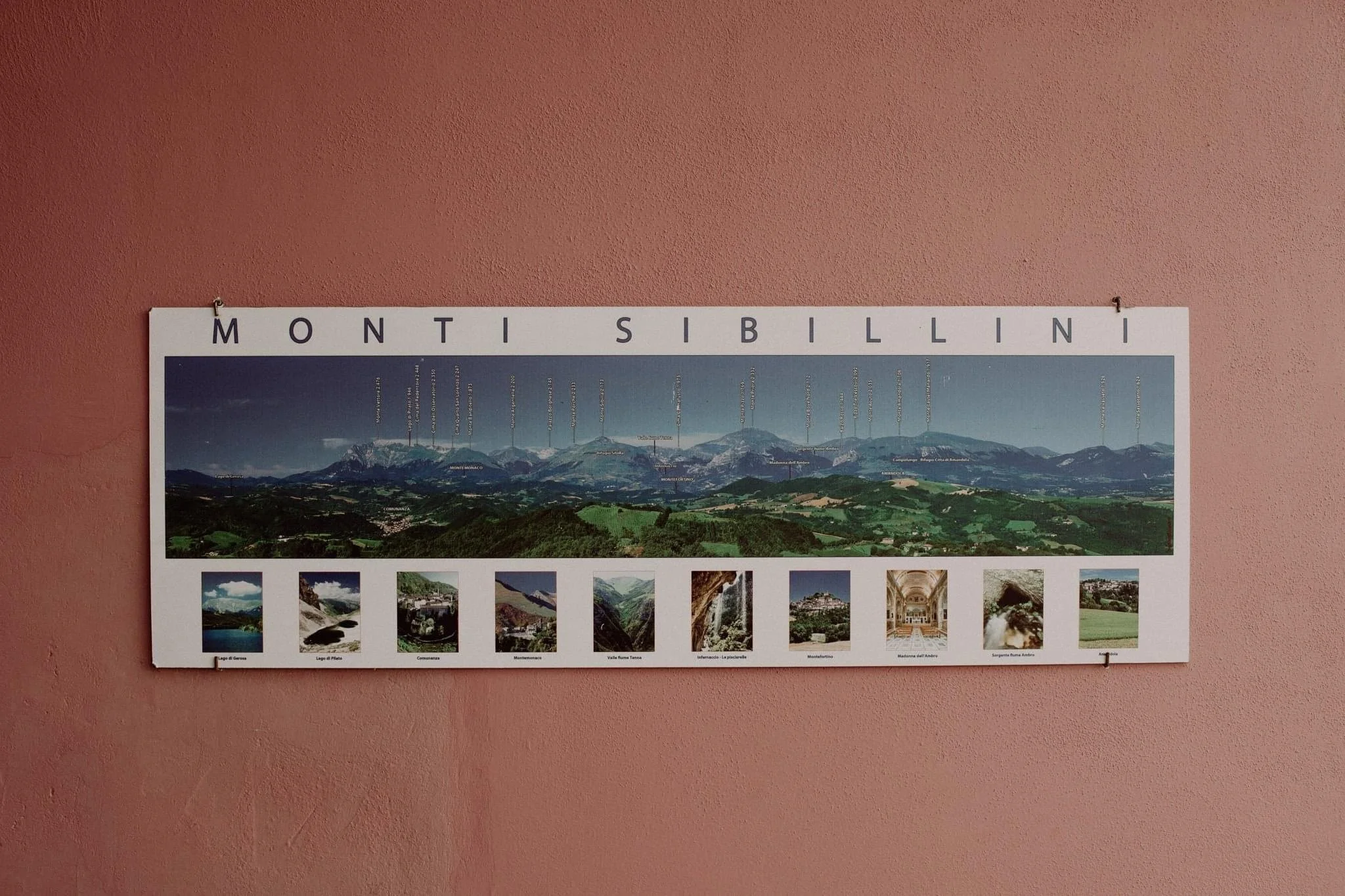 Panoramic informational poster of the Monti Sibillini mountain range displayed on a pink wall, with images and labels of notable locations and features.