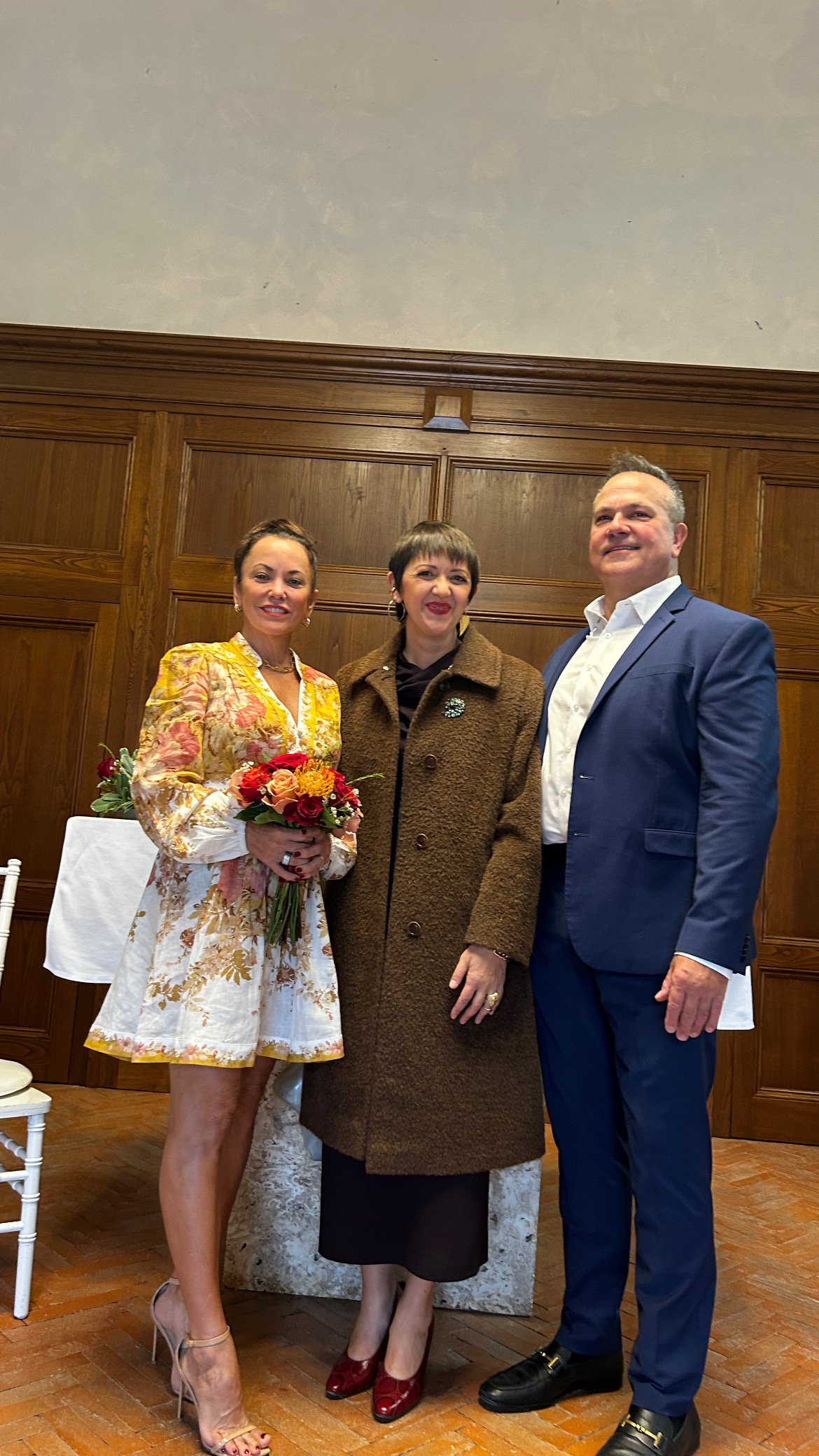 Three people standing together in a room with wooden paneling. The woman on the left is holding a bouquet of flowers, wearing a floral dress with high heels. The woman in the middle is wearing a brown coat, earrings, and high heels. The man on the ri