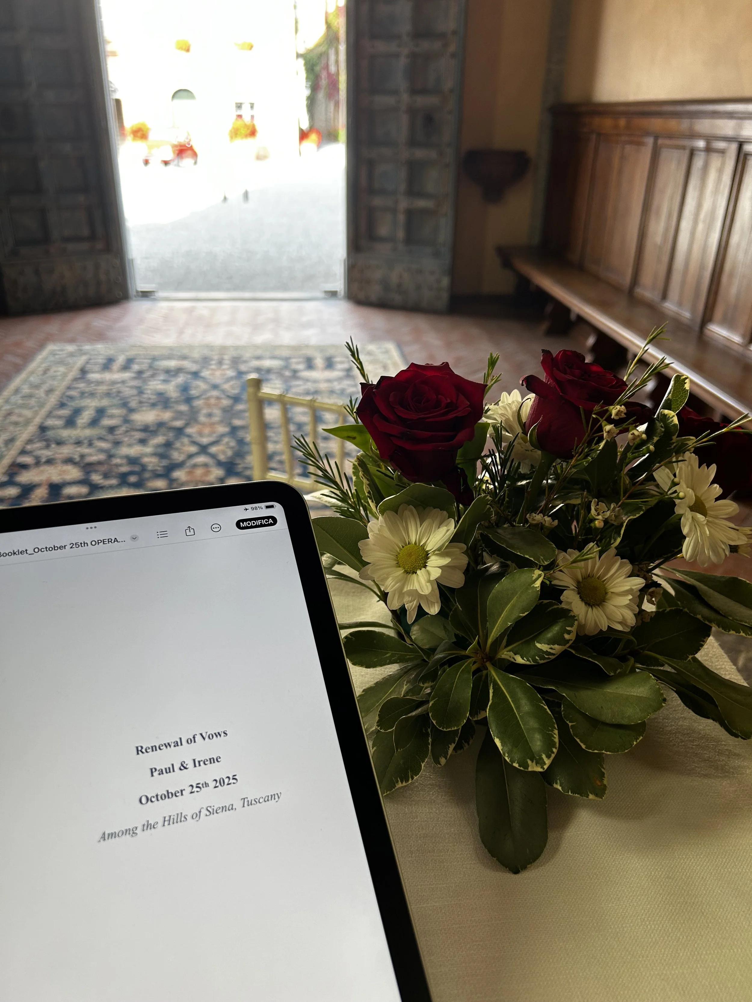 A tablet displaying a wedding vow document on a table with a floral arrangement of red roses and white daisies nearby, with a wooden bench inside a room leading to an open door outside.
