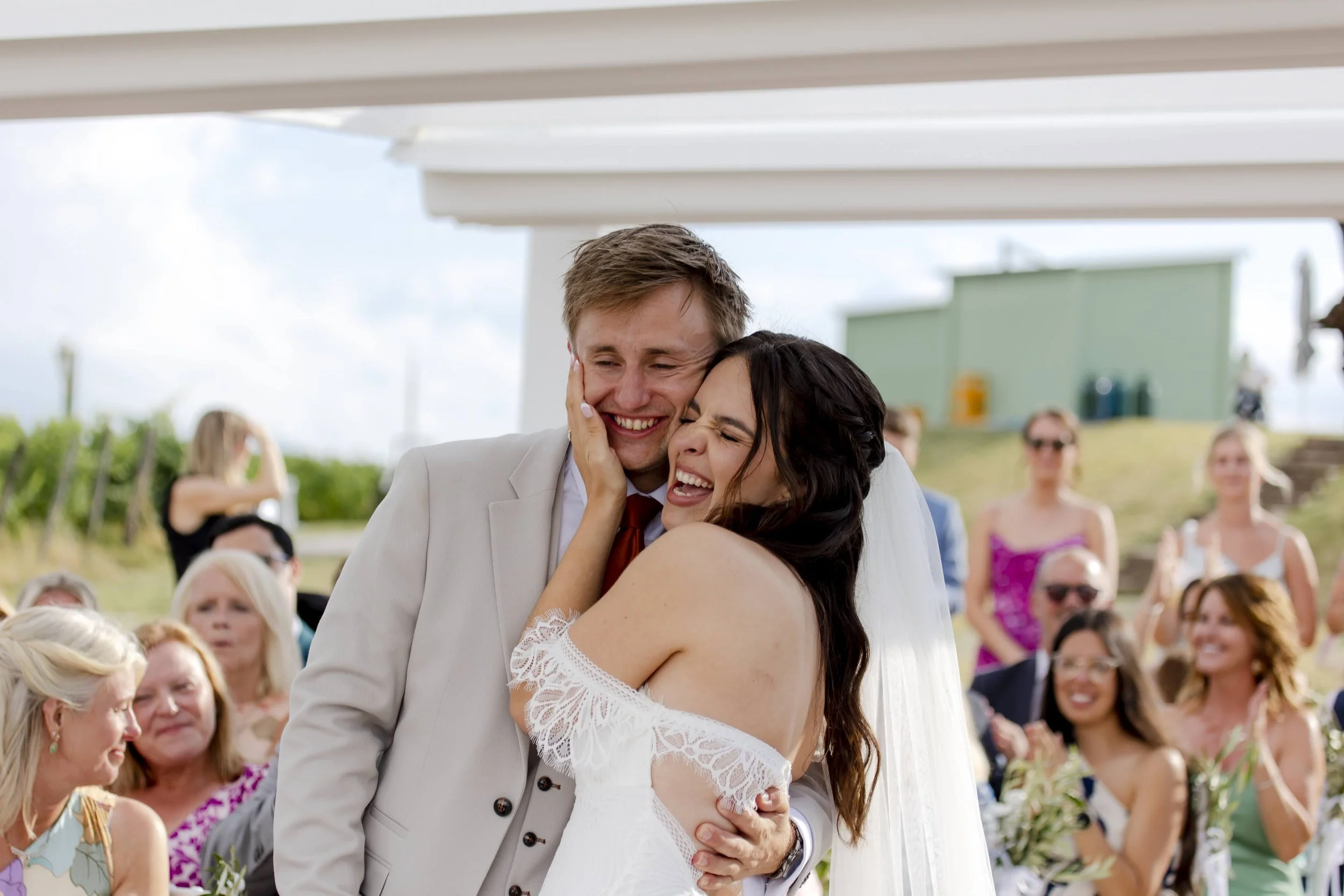A bride and groom sharing a joyful embrace at their outdoor wedding, surrounded by smiling guests.