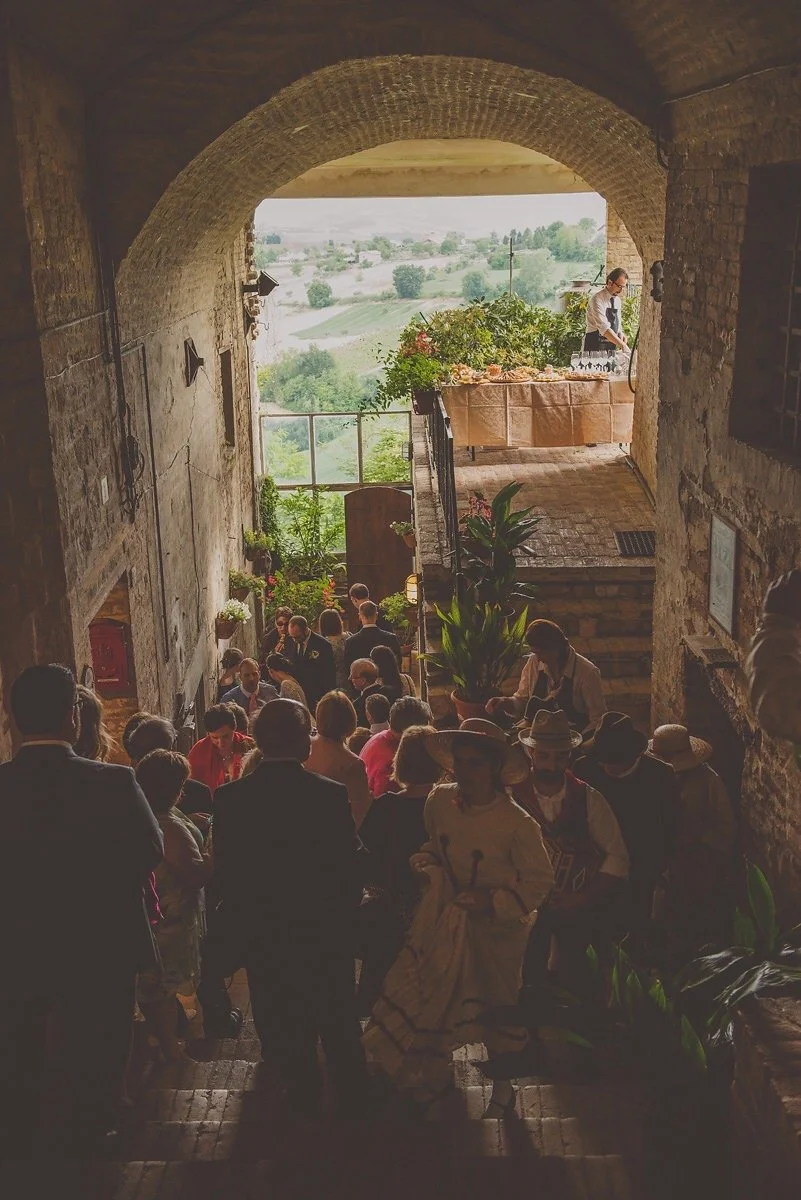 People gathered at a rustic indoor venue with a view of a countryside landscape, a woman preparing drinks behind a table, and plants decorating the space.