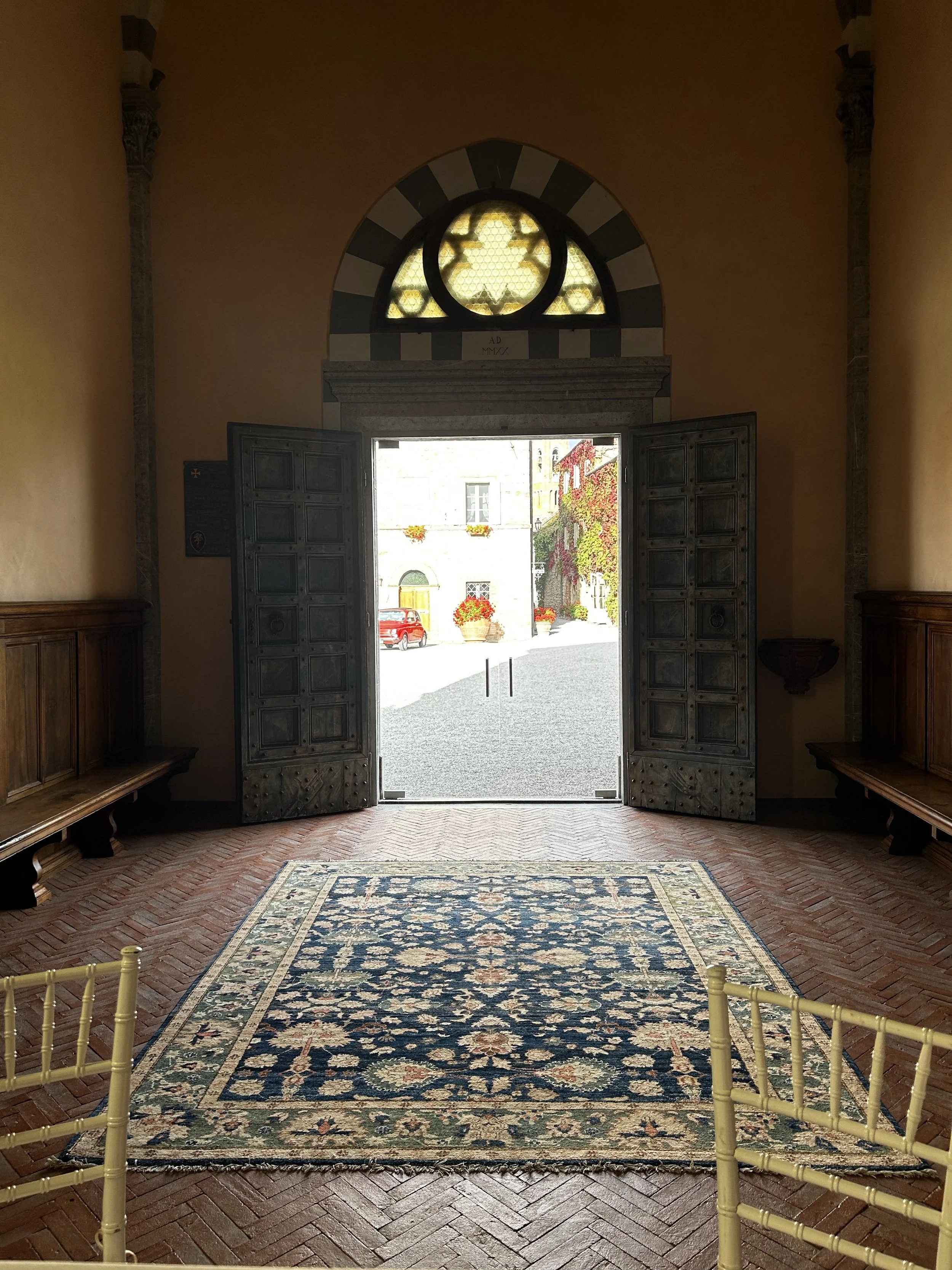 Open double doors leading outside to a cobblestone street with buildings and flower pots, sunlight streaming in, with a decorative carpet on the floor and wooden benches on sides.