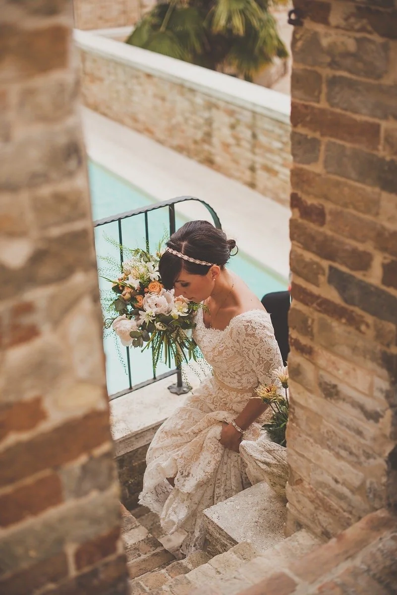A bride in a lace wedding dress sitting on stone stairs next to a brick wall, holding a bouquet of flowers, with her head bowed and eyes closed.