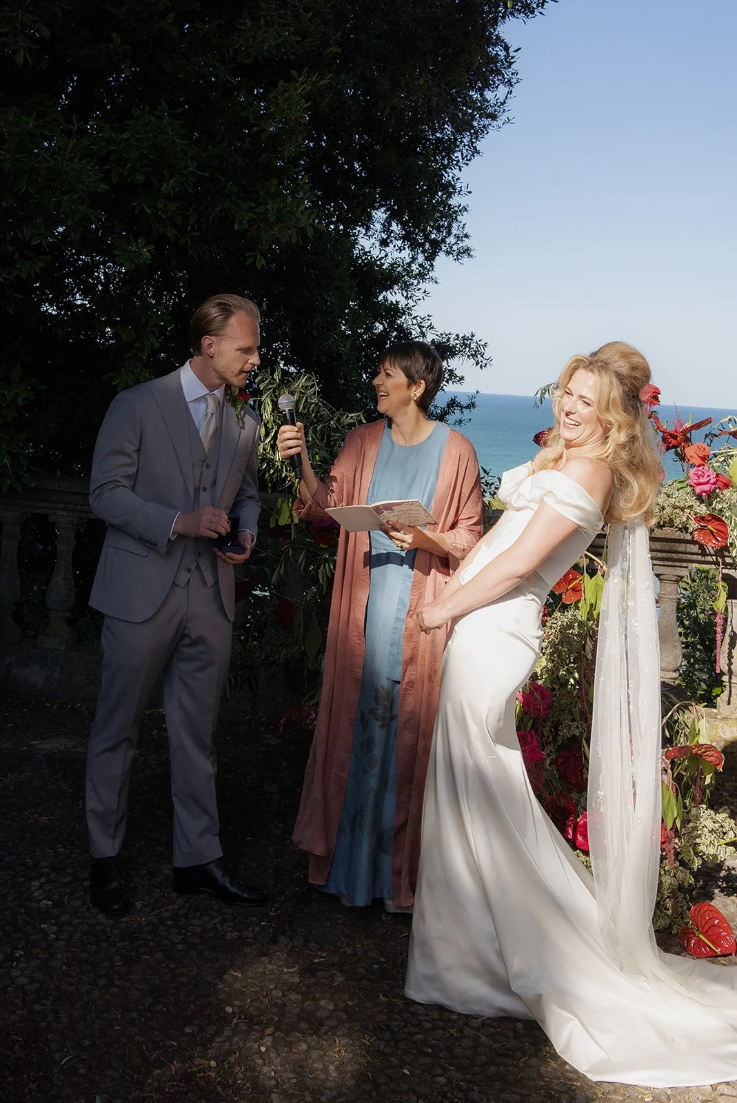 A wedding ceremony taking place outdoors with the ocean and sky in the background. The bride, wearing an off-shoulder white dress and veil, is smiling and holding her hands together. The groom is dressed in a light gray suit and tie, holding a small 