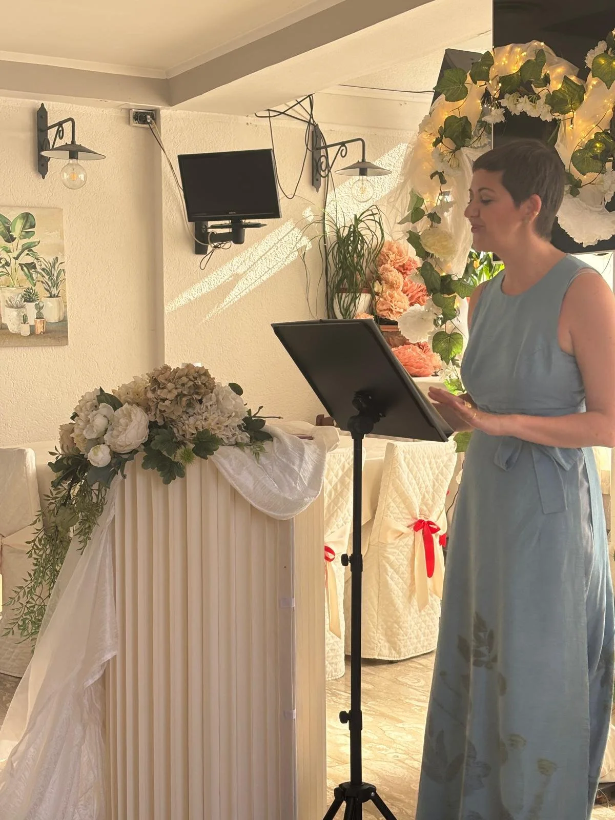 A woman with short dark hair in a sleeveless light blue dress standing near a floral arrangement, reading from a black tablet on a stand, with a decorative floral backdrop behind her, inside a room with cream crosses and wall lights.