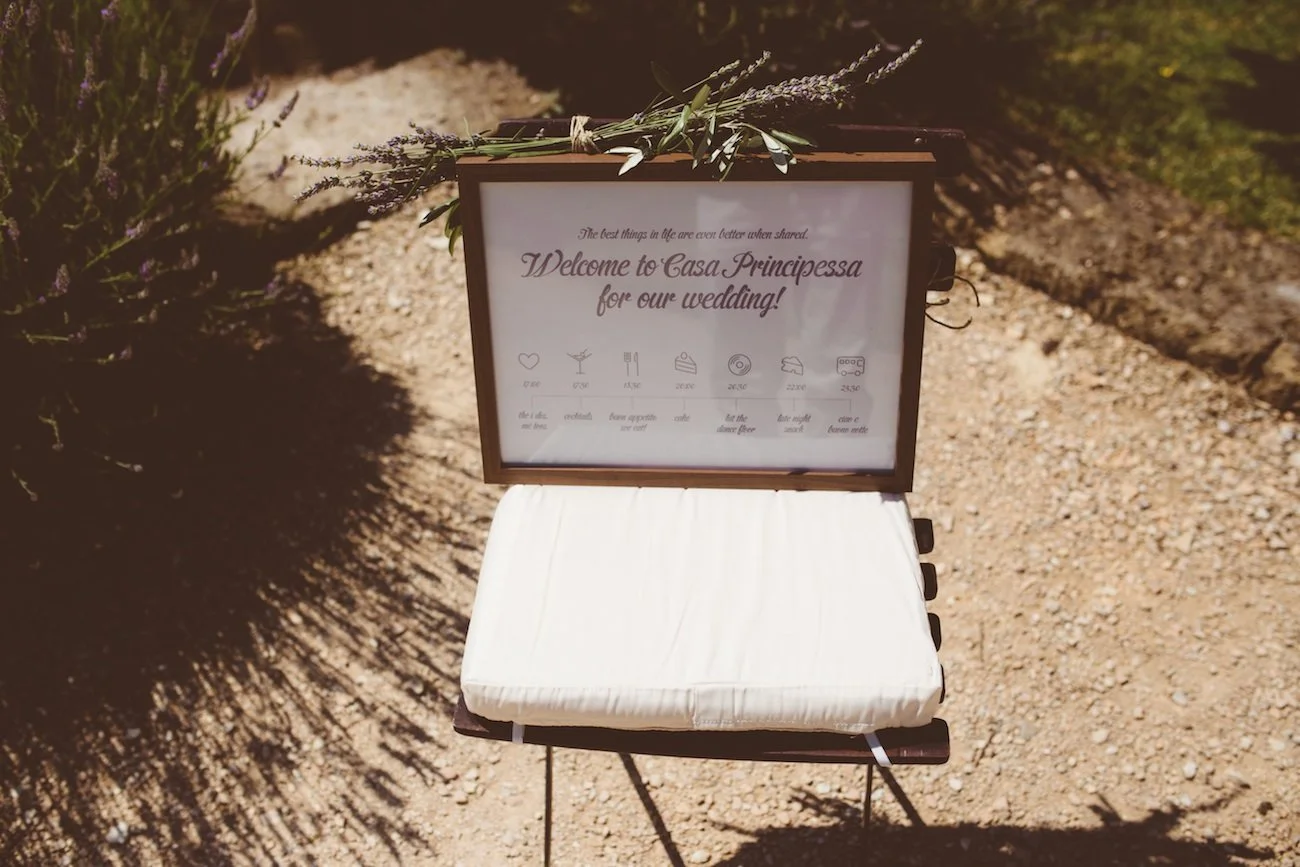 A wedding welcome sign on a stand with a cushion, surrounded by lavender flowers, on a dirt pathway.