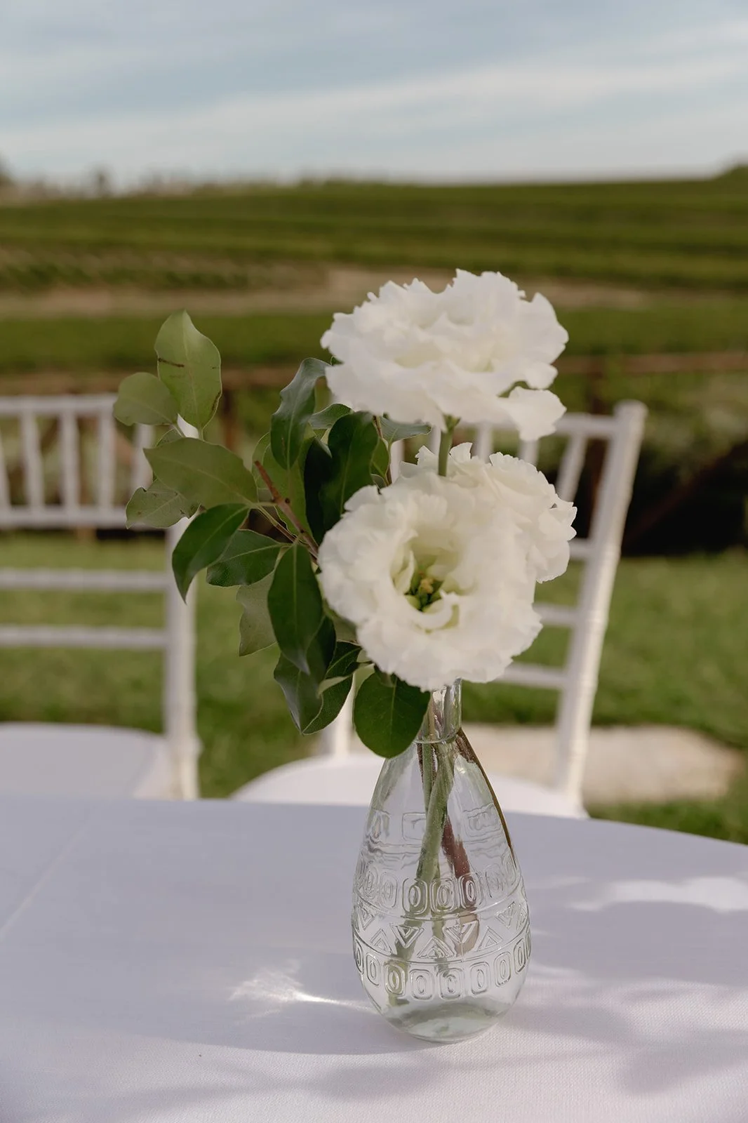 White flowers in a small glass vase on a white table outdoors, with chairs and a green landscape in the background.