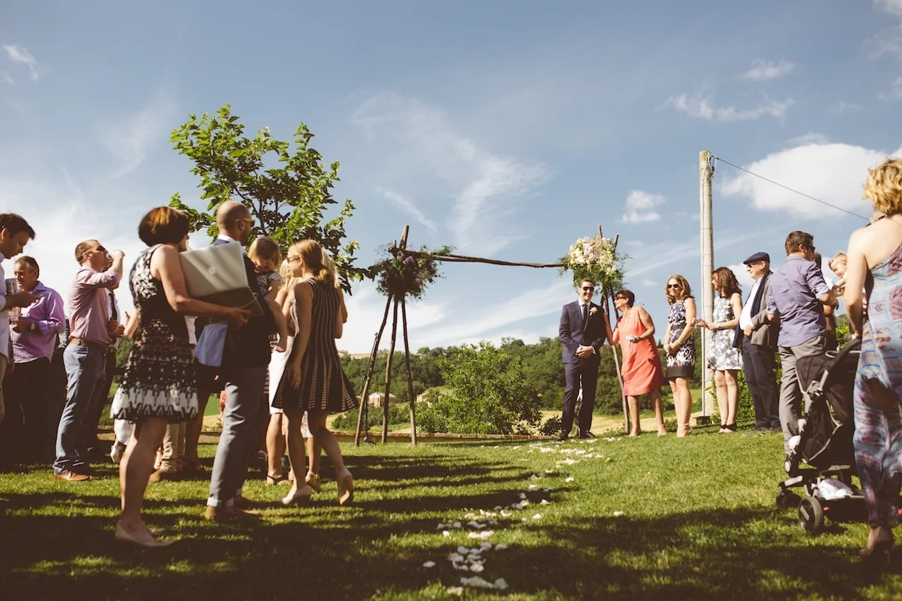 Guests gathered outdoors for a wedding ceremony beneath a wooden arch decorated with flowers, on a sunny day with a clear sky and trees in the background.