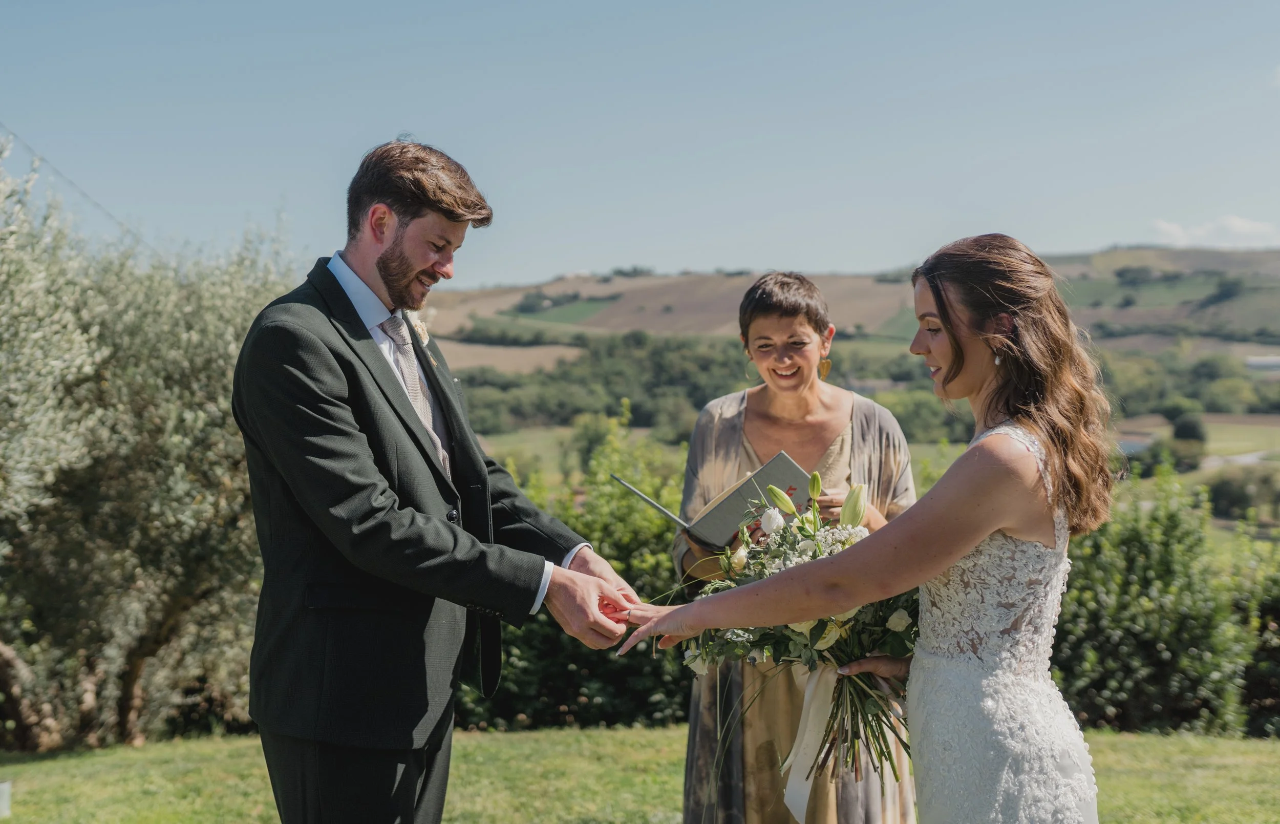 A couple getting married outdoors during a sunny day, with an officiant standing between them and smiling, holding a book. The bride is placing a ring on the groom's finger, both dressed in wedding attire, with a bouquet of flowers in her hand.