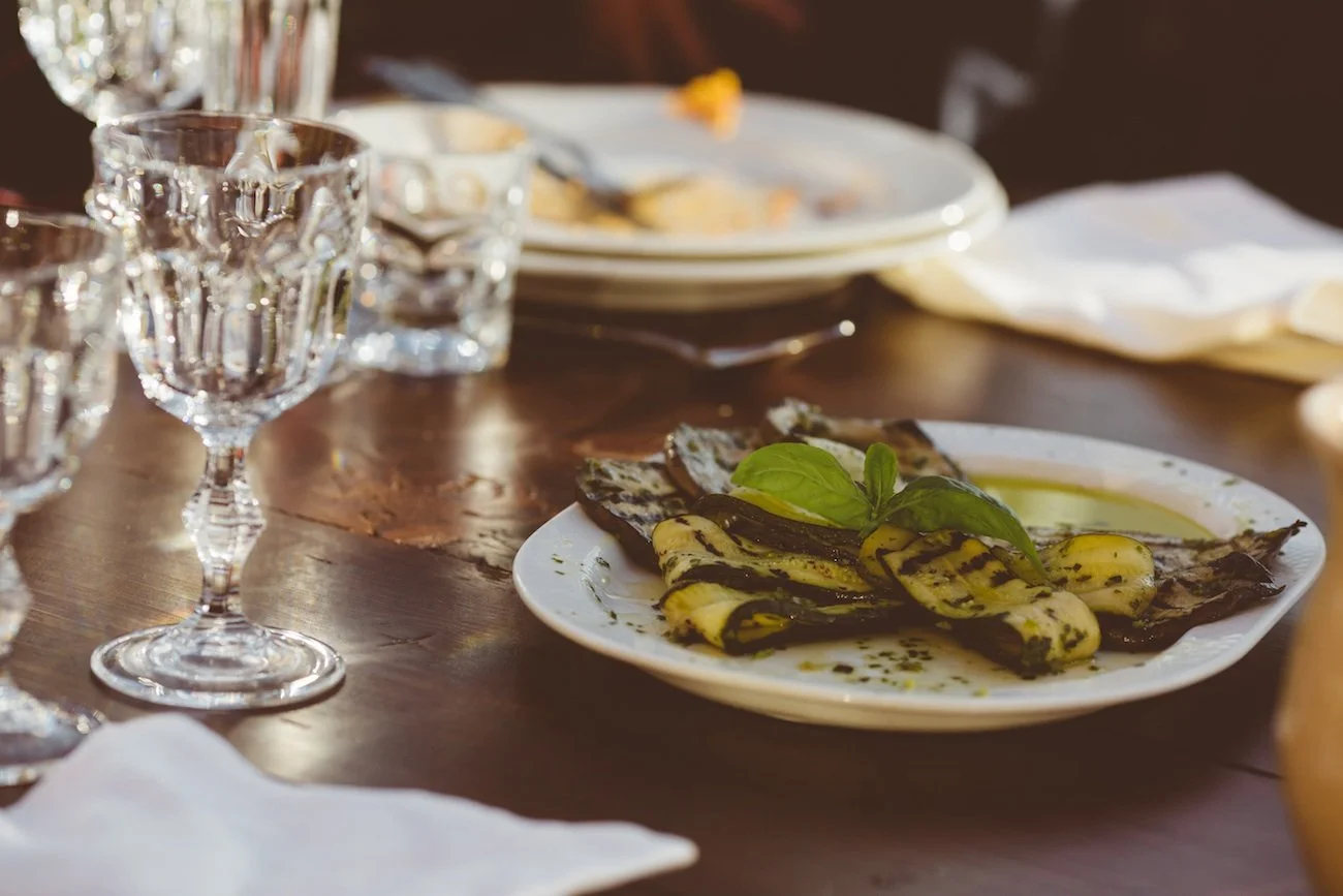 A plate of grilled bananas drizzled with chocolate and garnished with basil leaves on a wooden table, with empty glasses and plates in the background.