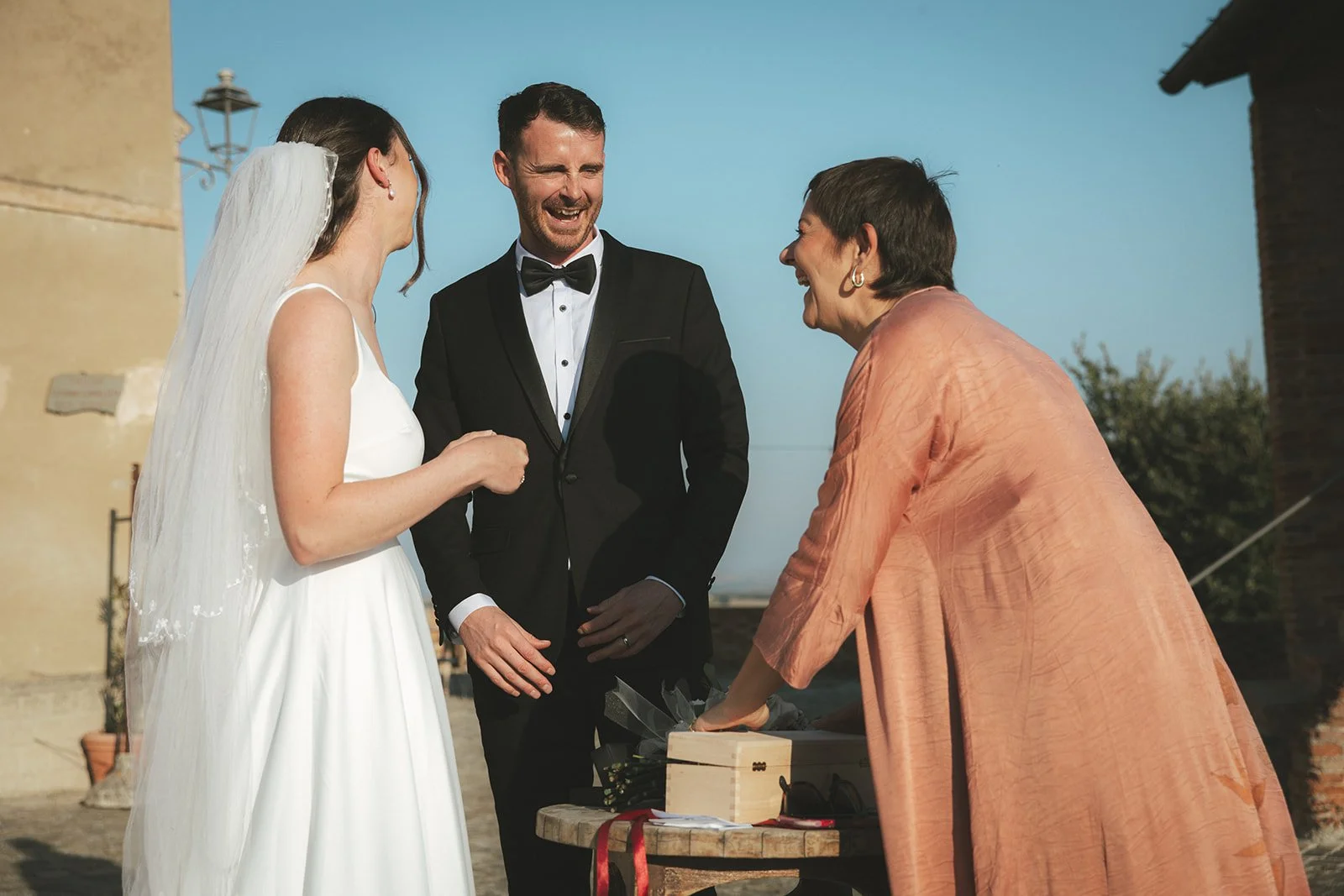 A bride and groom smiling and laughing during their wedding ceremony, with an officiant woman placing a gift or box on a table outside.