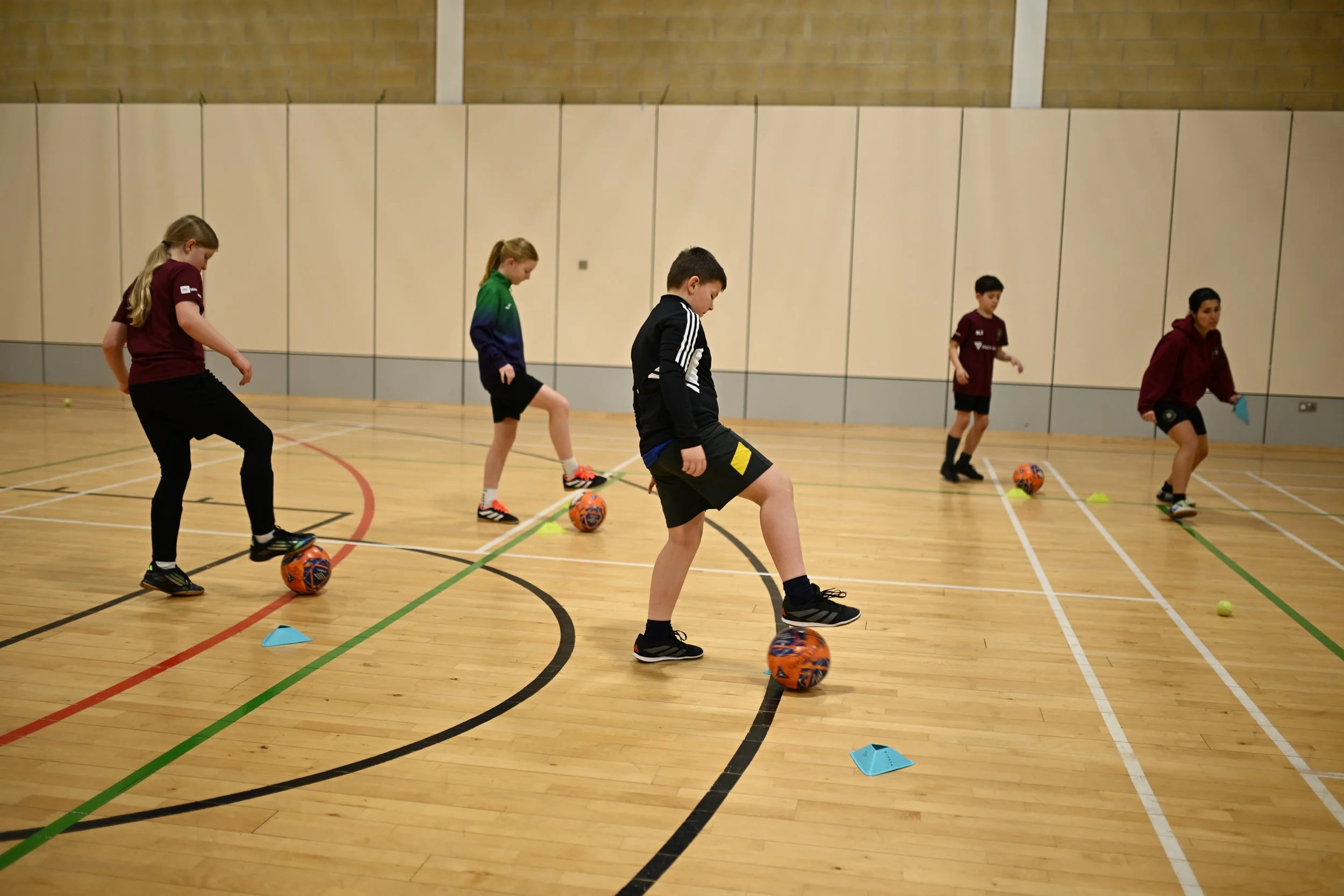 Children practicing soccer in an indoor gym, using orange soccer balls and small cones.