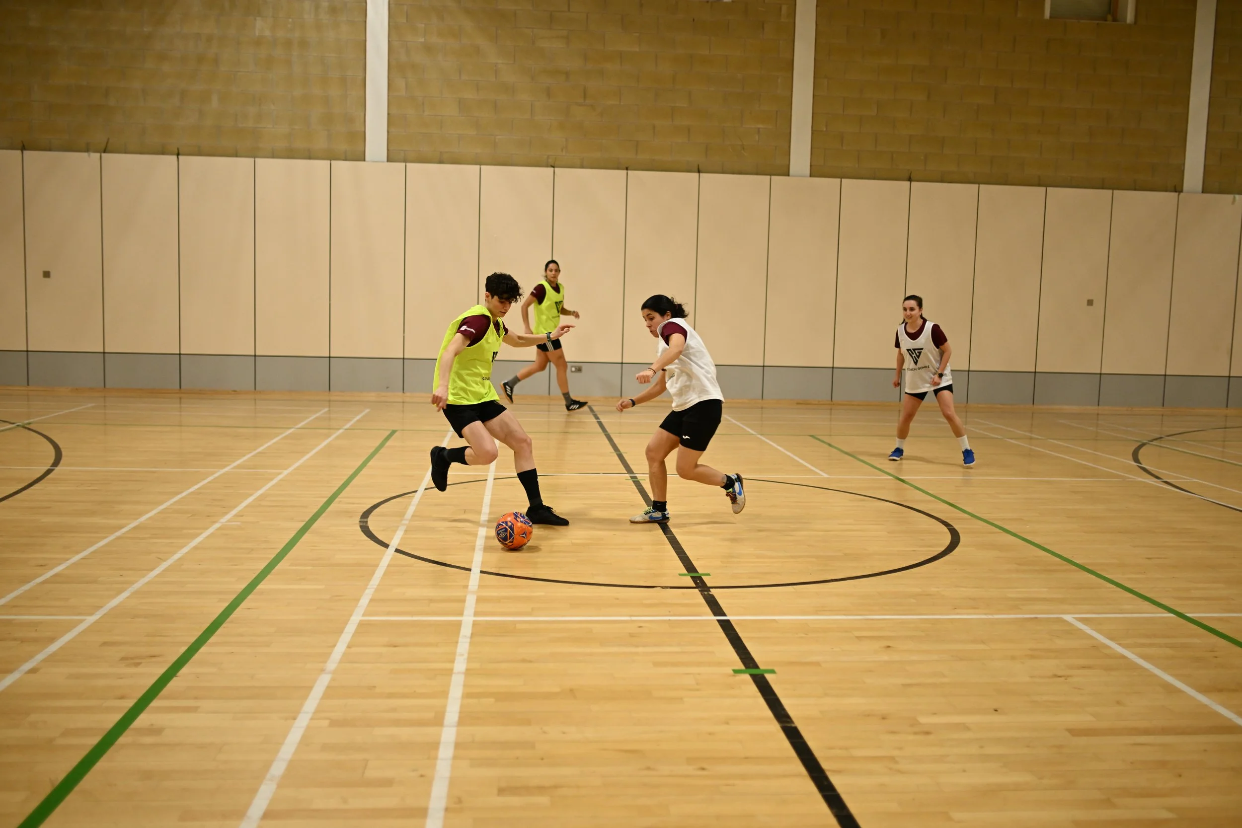 Four women playing indoor soccer in a gymnasium, two are actively engaged with the ball while the other two observe nearby.