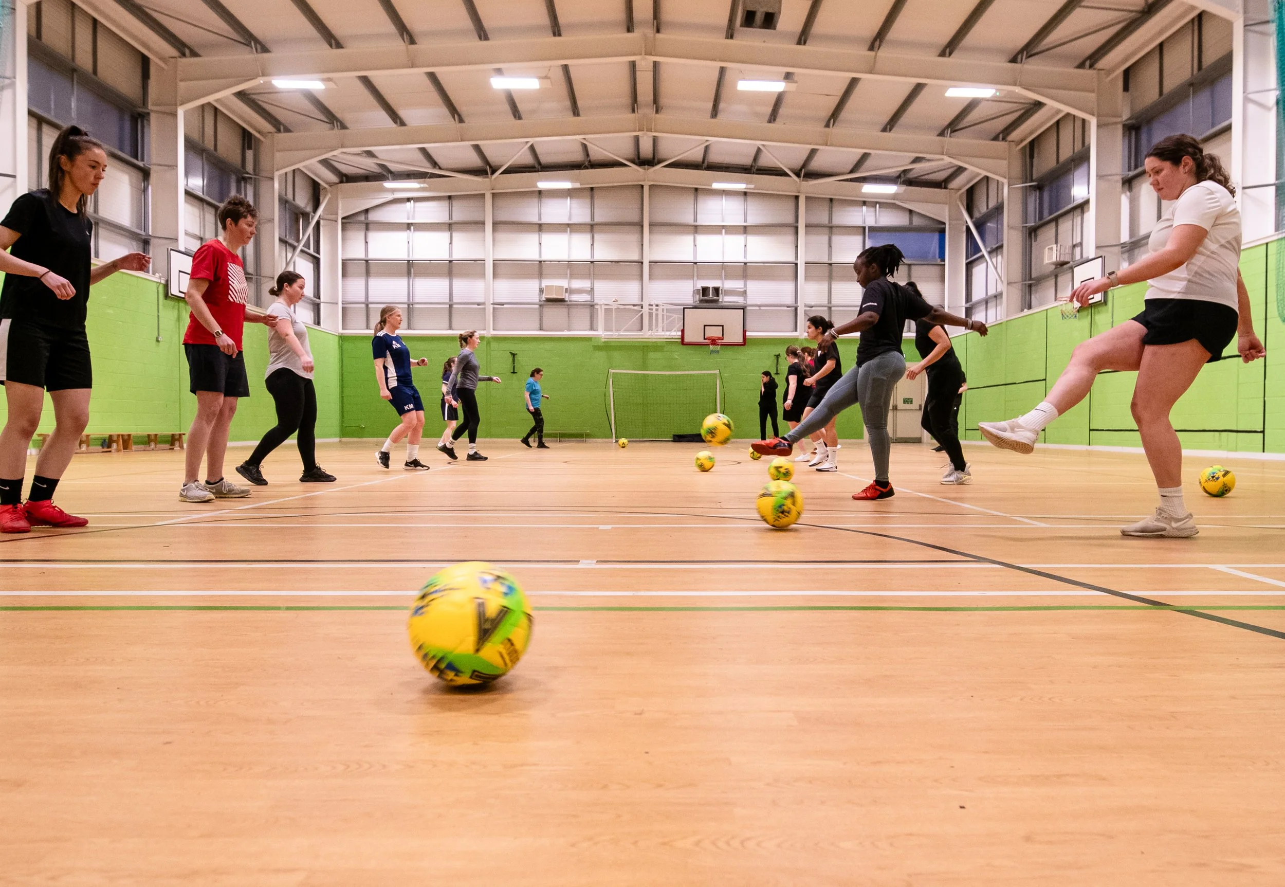 A group of people participating in a soccer practice inside an indoor gym with green walls and a wooden floor. They are performing drills with yellow soccer balls.