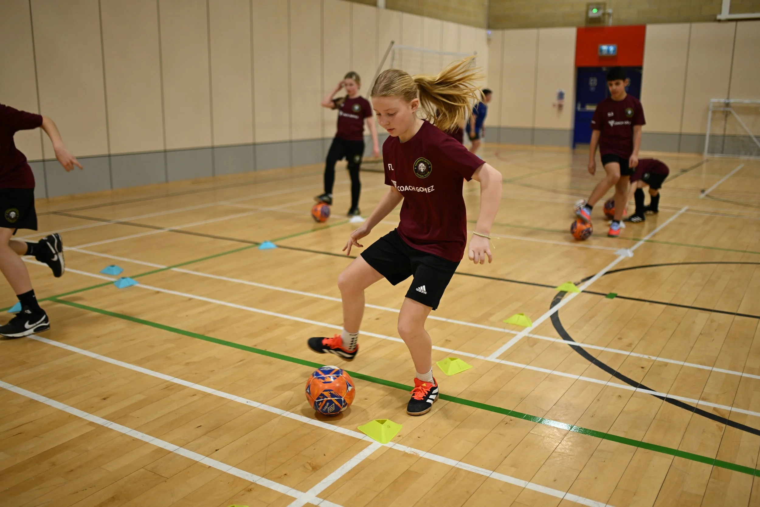 Children in a gym practicing soccer skills with balls and training cones.