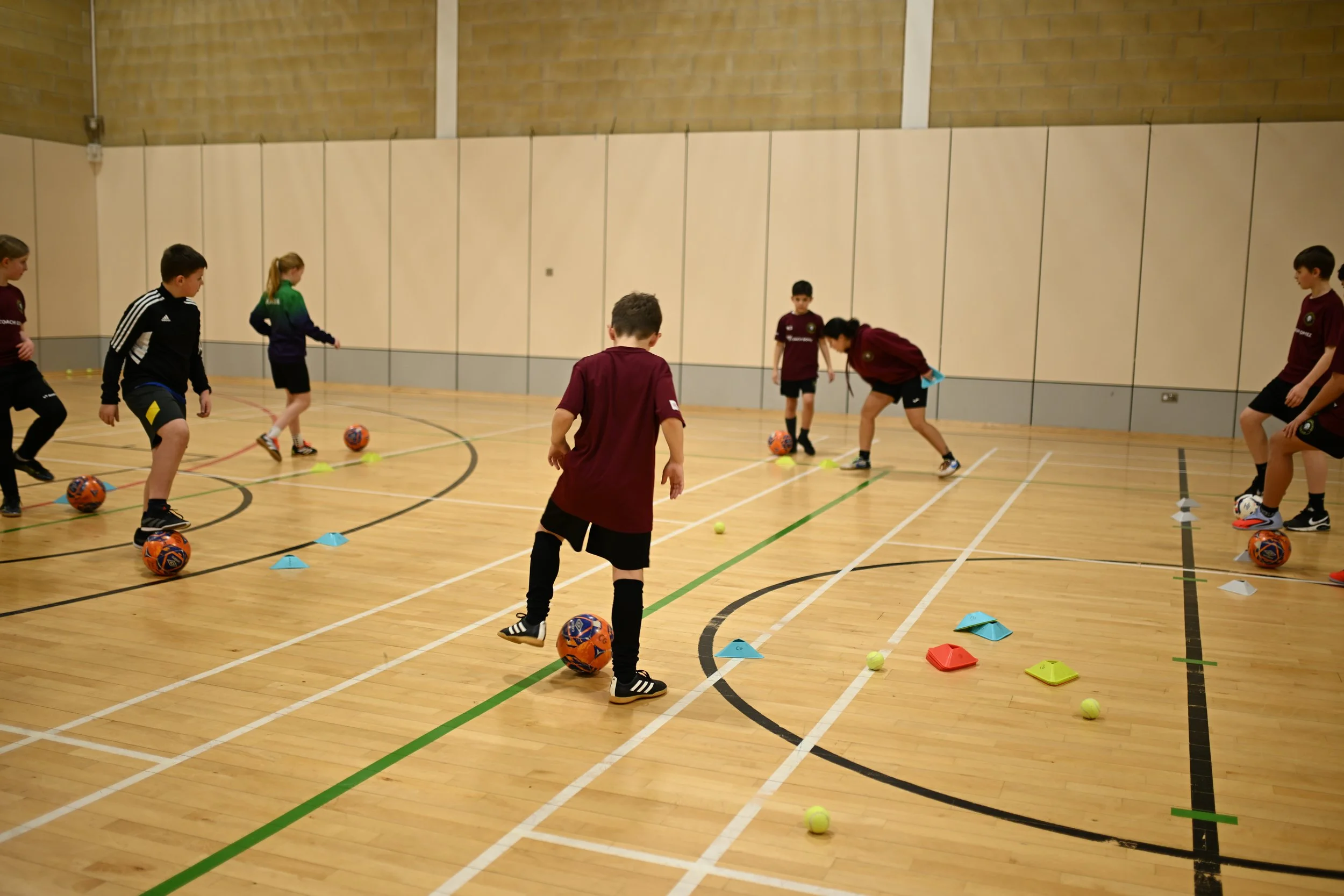 Children playing indoor soccer with balls and cones in a gymnasium.