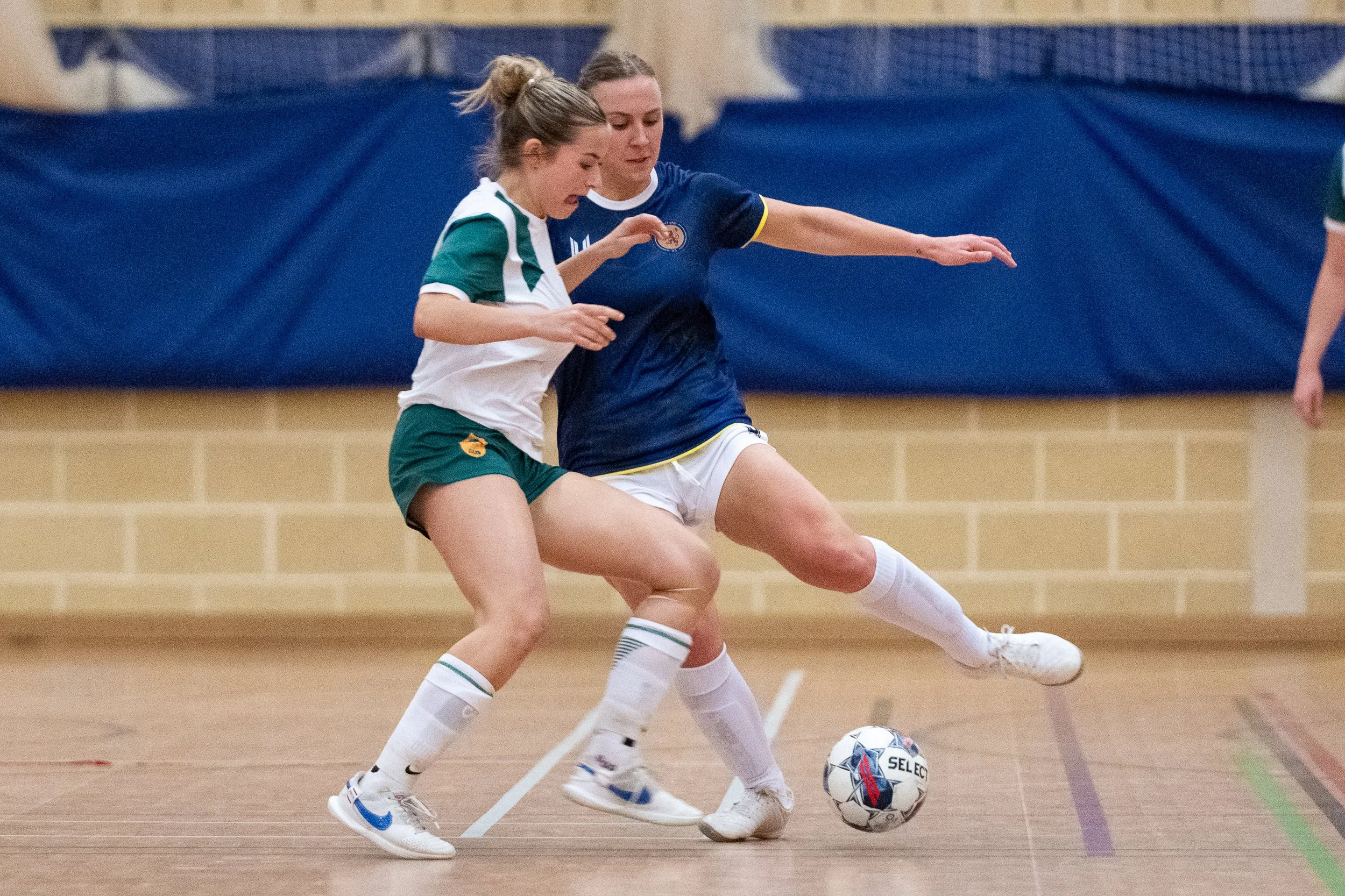 Two women playing indoor soccer, competing for the ball on the court.
