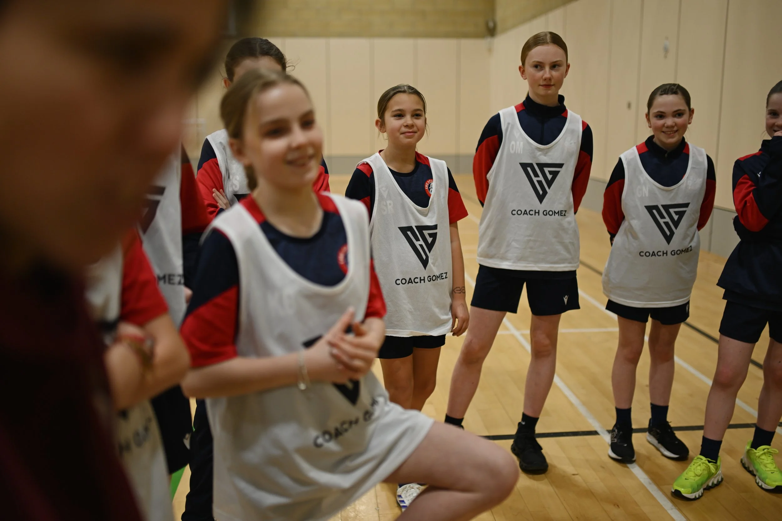 Young female basketball players standing in a gym, wearing white and red jerseys with 'Coach Gomez' printed on them, smiling and listening to their coach.