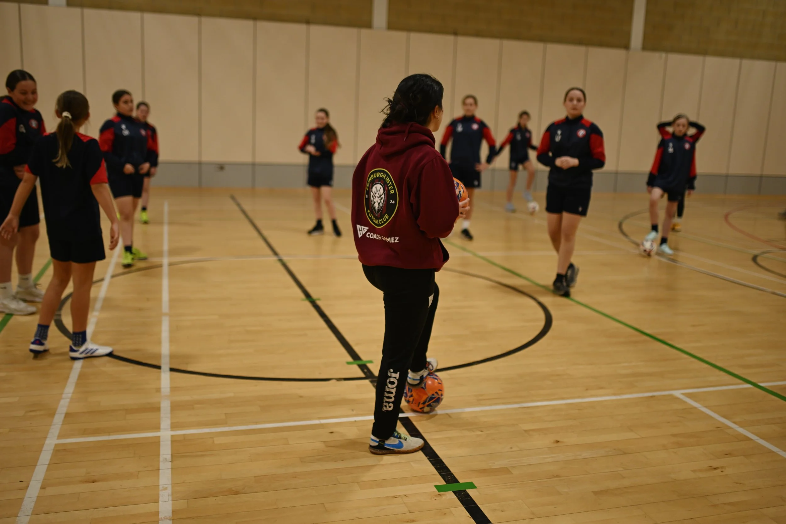 Woman coach in a maroon hoodie stands on a gym floor with a soccer ball at her feet, observing a group of young girls in sports attire during indoor soccer practice.