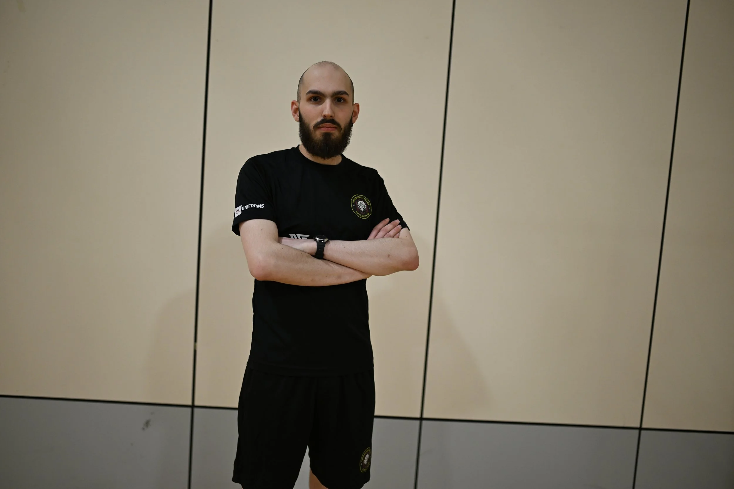 A man with a beard and bald head standing with arms crossed in front of a beige wall with black lines.