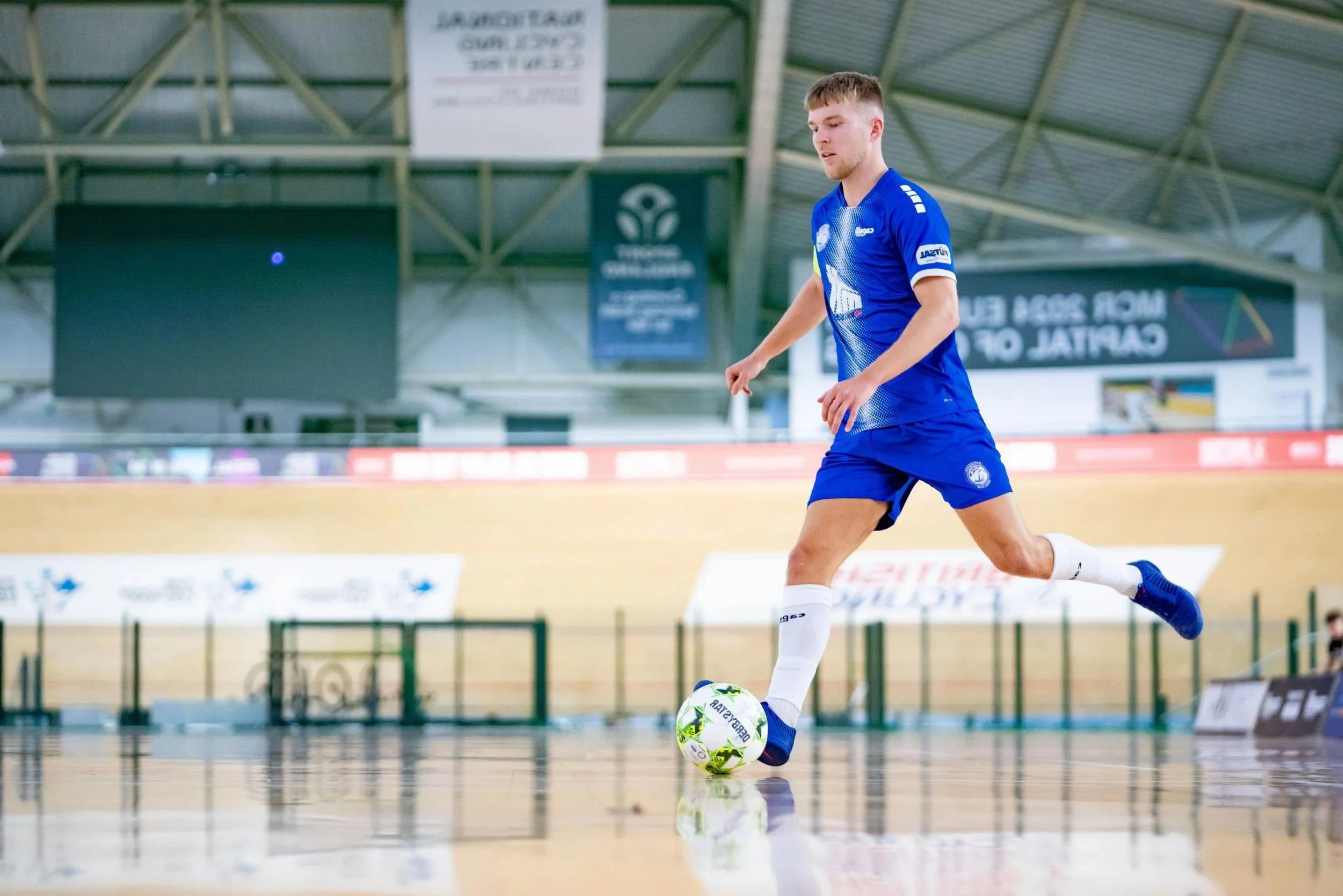 A male soccer player in a blue uniform kicks a soccer ball on an indoor field, with banners and a scoreboard in the background.